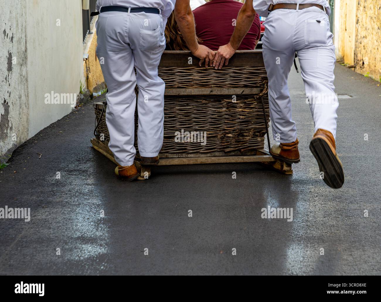 Due uomini in uniforme bianca spingono un toboga in vimini lungo una strada stretta, mostrando un metodo di trasporto tradizionale Funchal Madeira Portogallo Foto Stock
