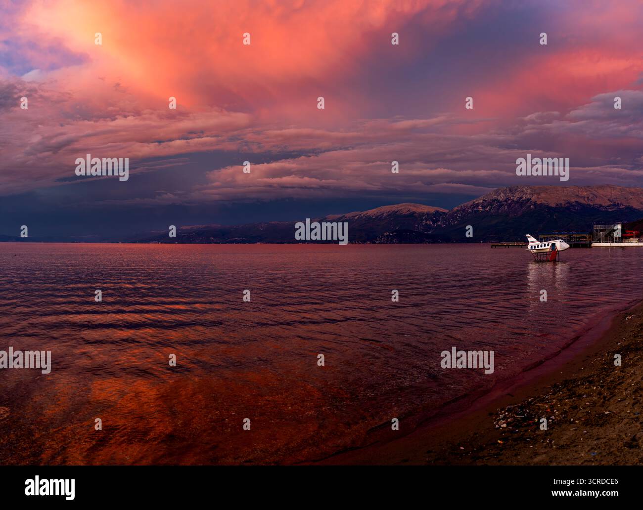 Spettacolare tramonto rosso sul lago di Ocrida a Pogradec, Albania, con montagne, nuvole luccicanti e una barca sulla riva Foto Stock