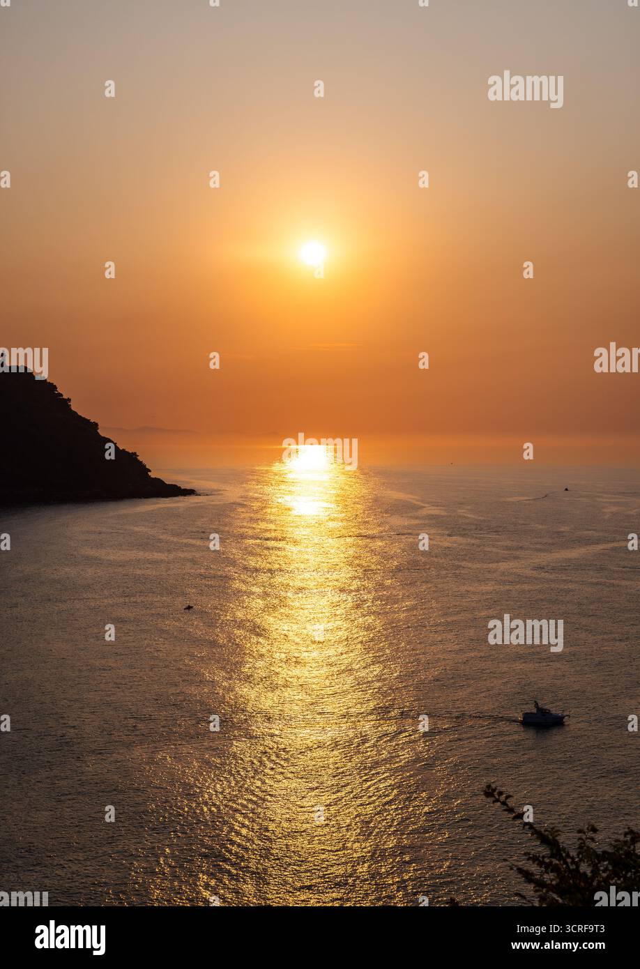 Vista del sole che tramonta sul mare tranquillo, proiettando un bagliore dorato sull'acqua e sagomando la costa rocciosa di San Sebastian, Donostia, Spagna. Foto Stock