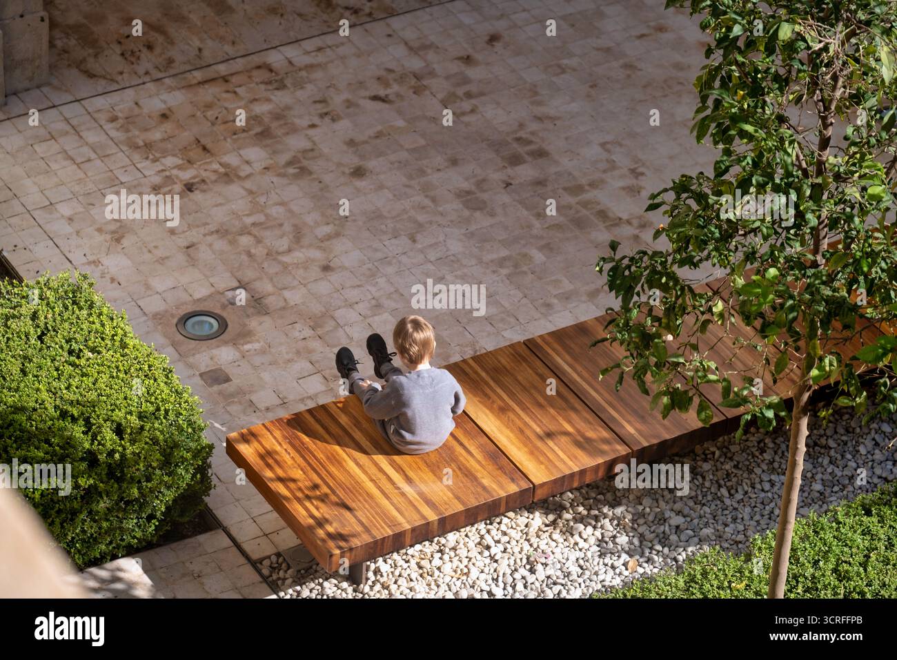L'immagine presenta una vista dall'alto di un ambiente esterno con una persona seduta su una panchina di legno. La persona sembra indossare un coagulo di colore chiaro Foto Stock