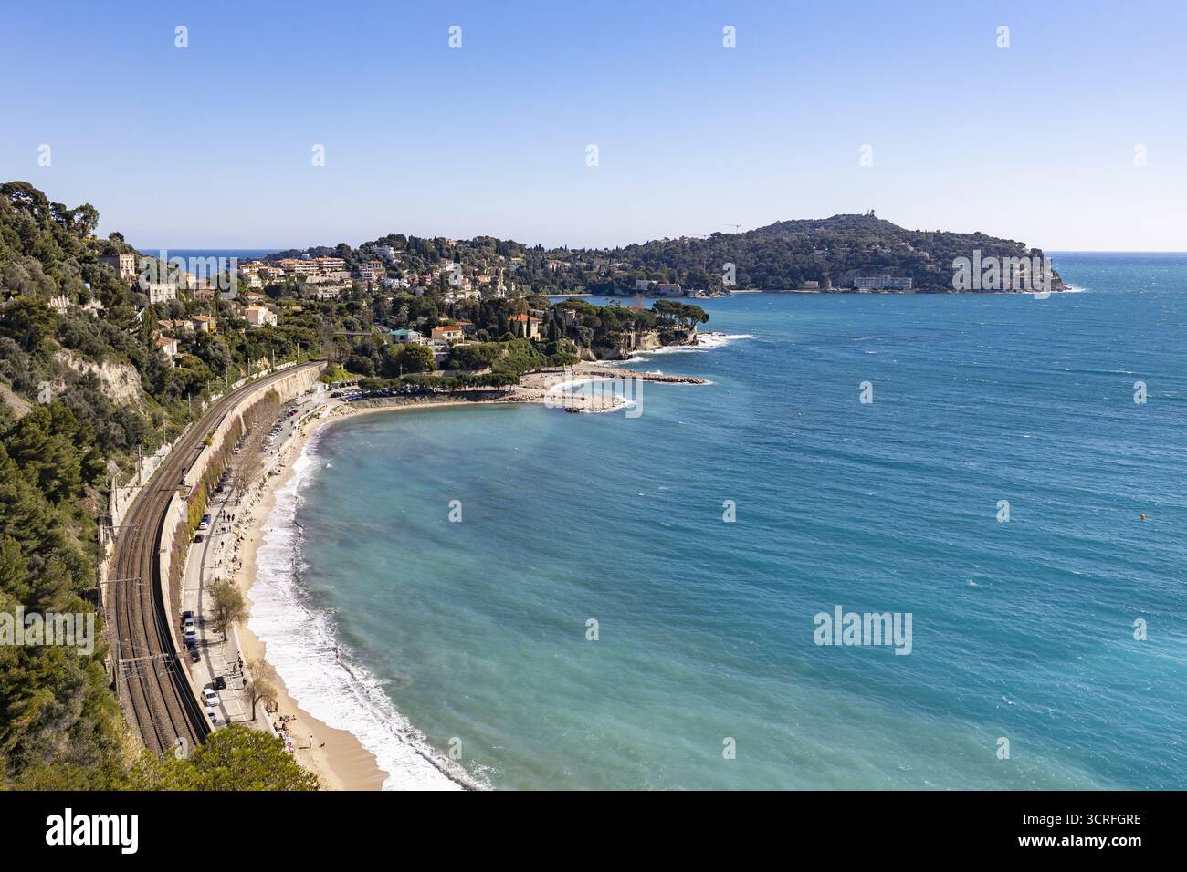 Vista di una vivace scena costiera con acque turchesi che incontrano una spiaggia sabbiosa, incorniciata da una vegetazione lussureggiante e una ferrovia tortuosa, Villefranche-sur-Mer, Prov Foto Stock