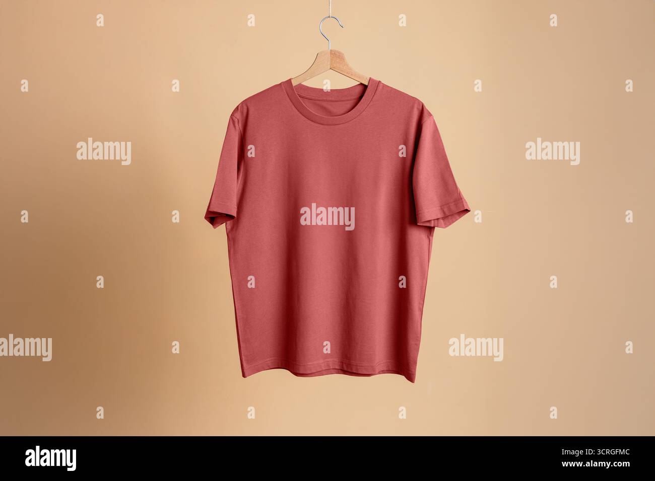 T-shirt in cotone premium (rosso terracotta/corallo) appesa su un gancio di legno. Mockup isolato su sfondo blu neutro. Ampio spazio di copia. Foto Stock