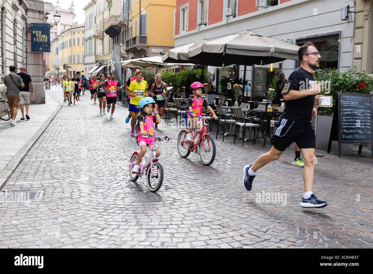 I corridori maratonesi avanzano sulle strade di ciottoli della città, mentre i bambini in bicicletta partecipano mettendo in risalto lo spirito della comunità familiare e gli sport urbani ener Foto Stock