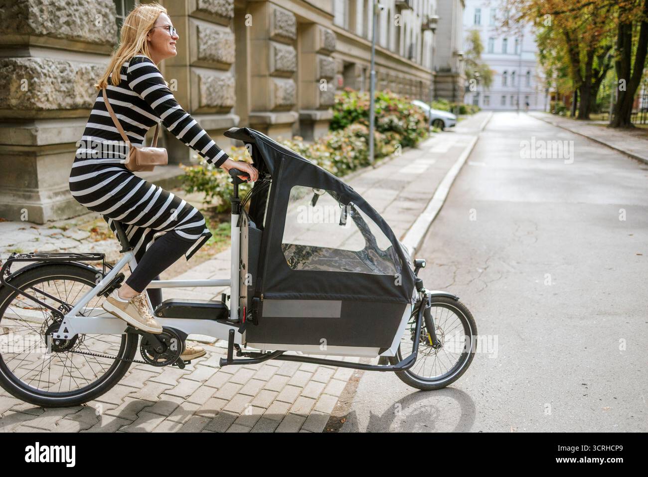 Giovane donna in sella a una bici cargo in città in una giornata di sole Foto Stock
