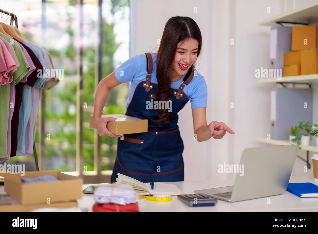Allegra imprenditrice asiatica elabora gli ordini di abbigliamento. Organizza l'inventario prepara le spedizioni utilizzando laptop per la gestione delle vendite online. SMA Foto Stock