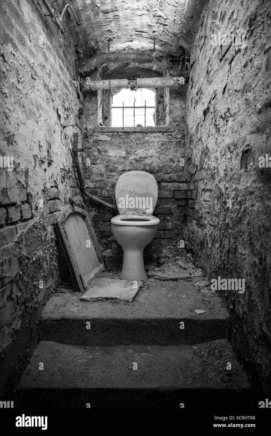 Toilette abbandonata nell'edificio in decadenza con pareti incrinate e luce naturale attraverso Rusty Window - Urban Exploration Art Foto Stock
