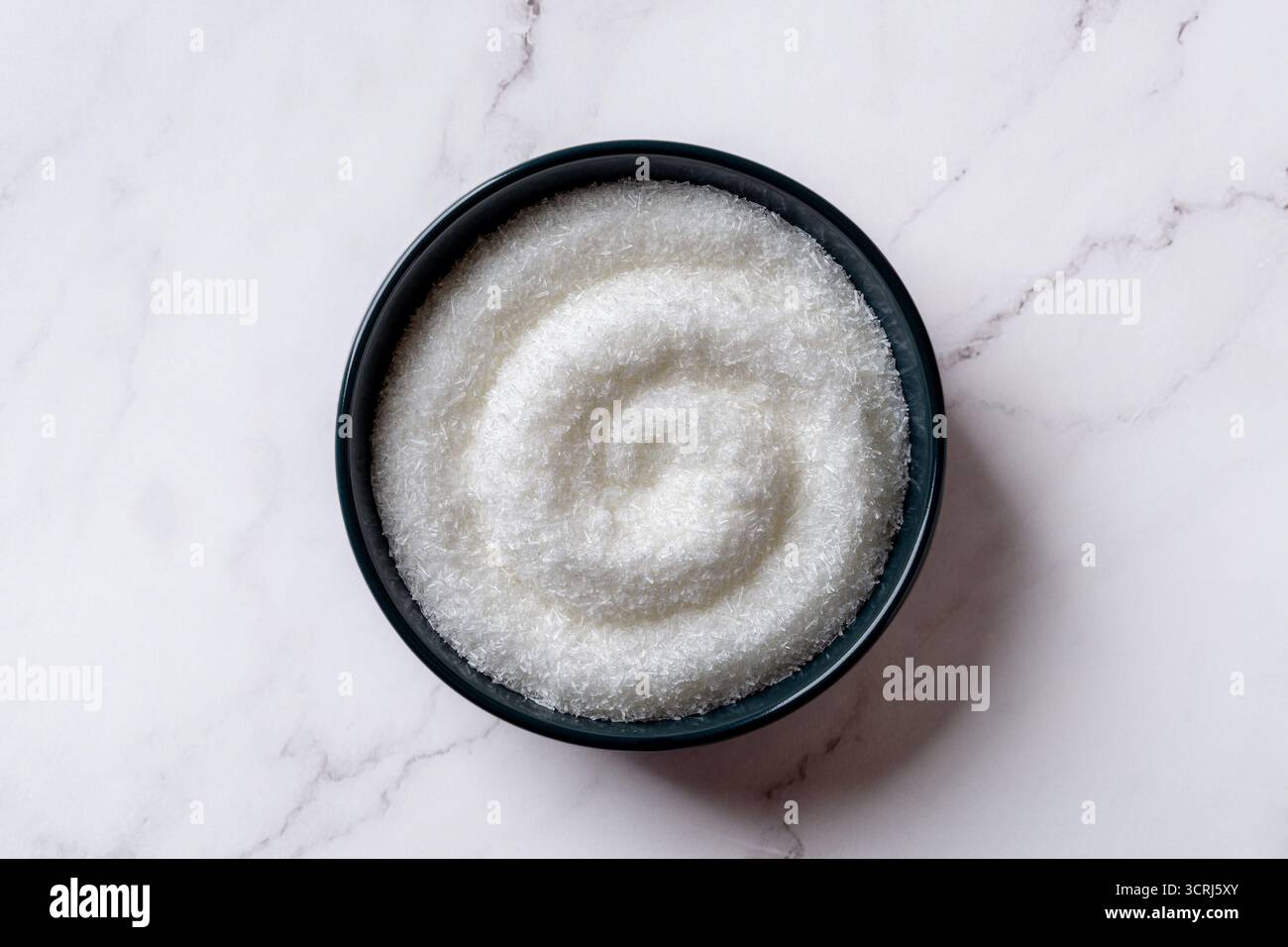 Glutammato monosodico in una ciotola blu scuro sopra il bancone della cucina in marmo. Piatto pieno di cristalli bianchi MSG. Concetto di additivo alimentare per esaltatori di sapidità E621. Foto Stock