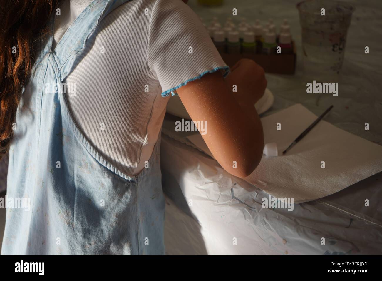 La ragazza dipinge a tavola, circondata da provviste. La luce del sole mette in risalto l'energia creativa pratica per l'apprendimento back-to-School. Foto Stock