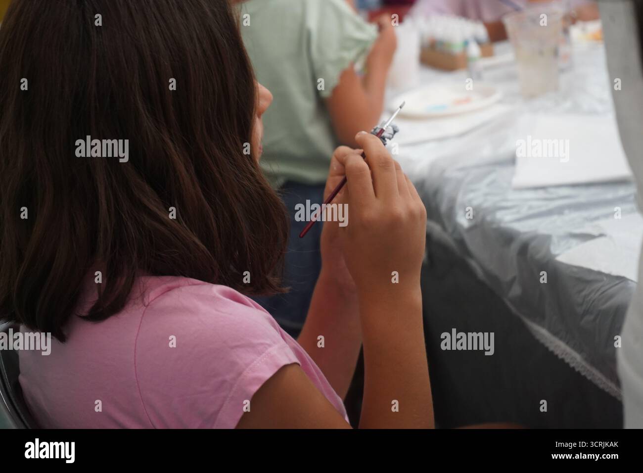 Una bambina di dieci anni con i capelli castani si concentra su un'attività artistica, tenendo in mano un pennello per dipingere una miniatura. Foto Stock