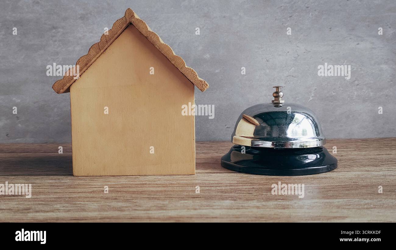 Campanello accanto al modello di casa in legno che simboleggia il servizio domestico, la richiesta di informazioni sulla proprietà e il supporto immobiliare Foto Stock