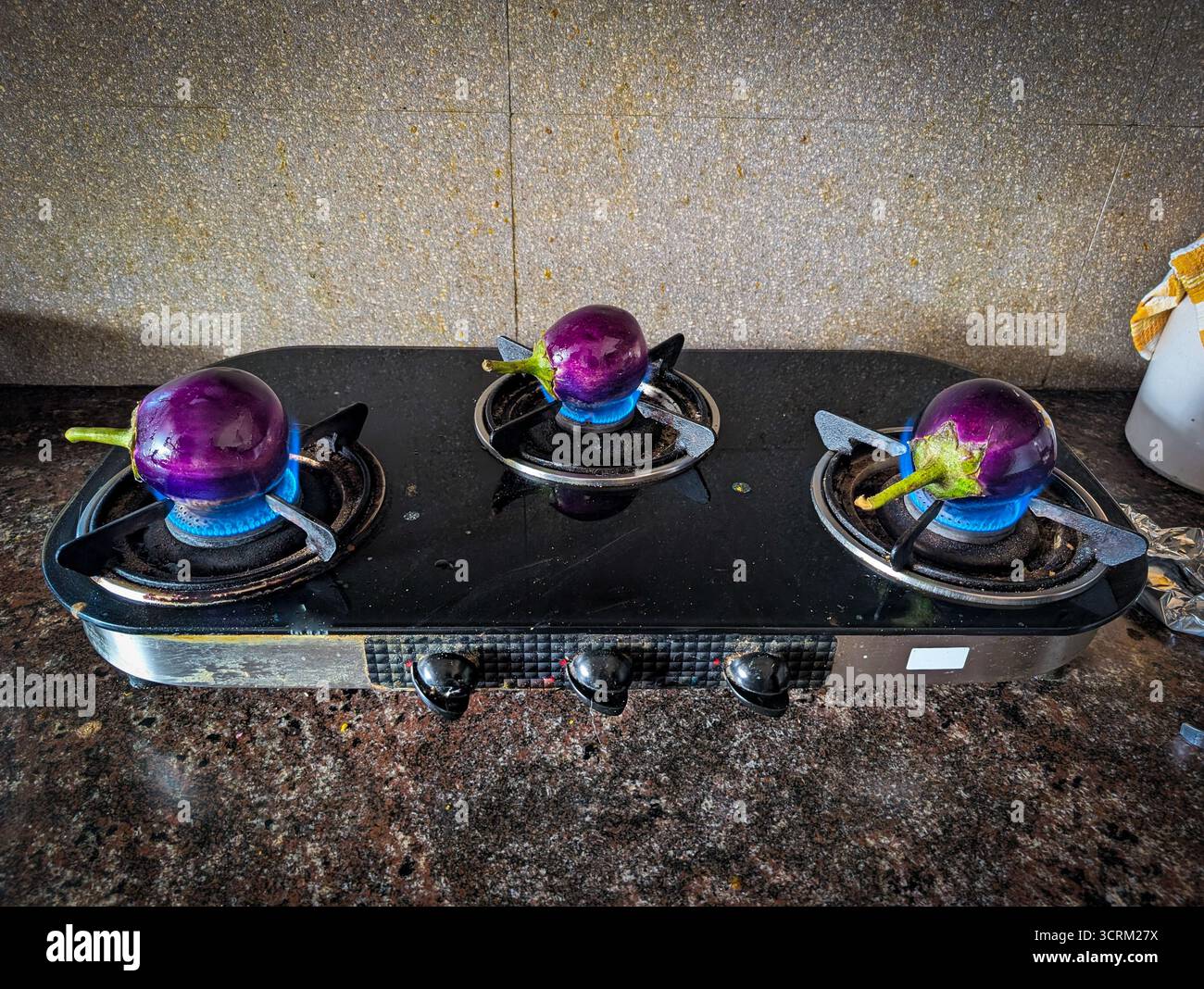 Tre melanzane viola arrostite su una stufa a gas con fiamme blu. Foto Stock