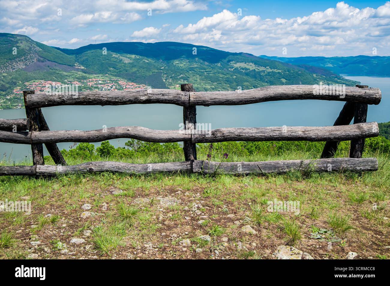 Una recinzione in legno si affaccia su un paesaggio panoramico con montagne e un lago sotto un cielo blu con soffici nuvole. Foto Stock
