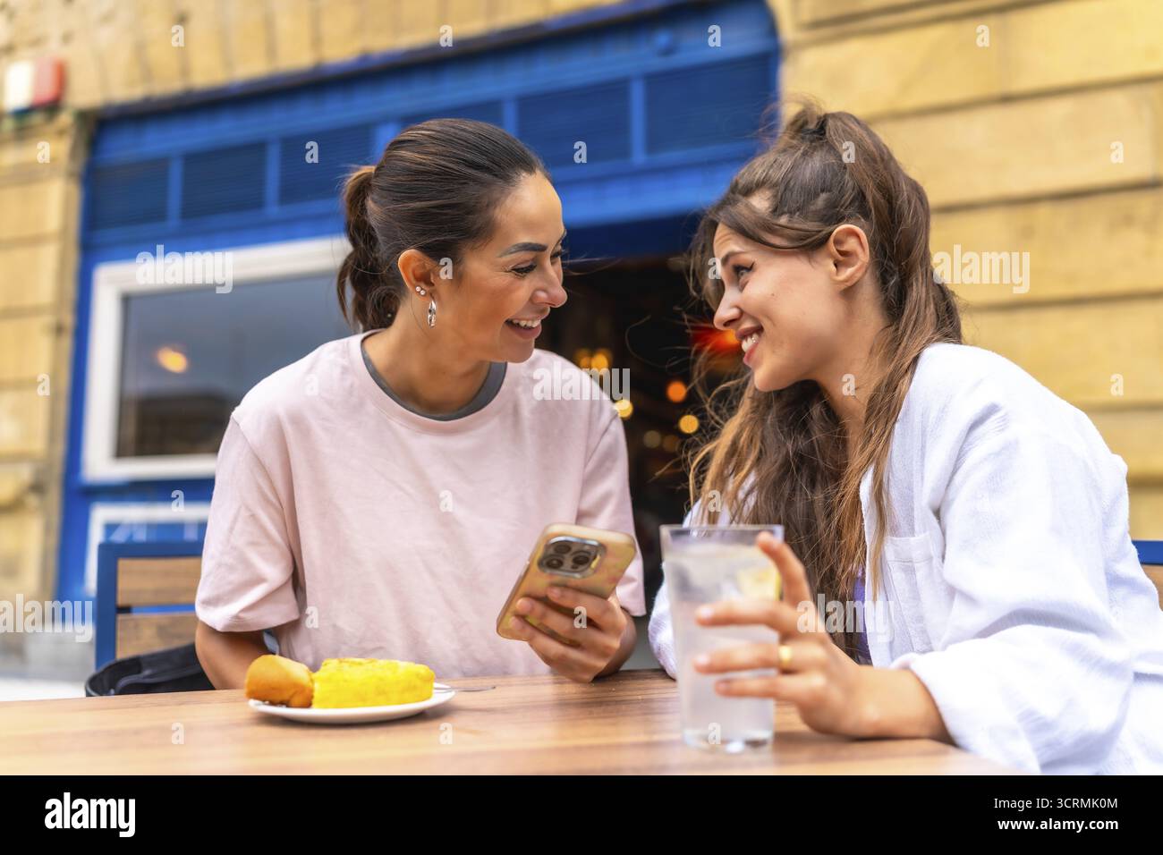 Due donne stanno gustando un pasto e un drink in un bar all'aperto, condividendo un momento di connessione mentre navigano insieme su uno smartphone Foto Stock