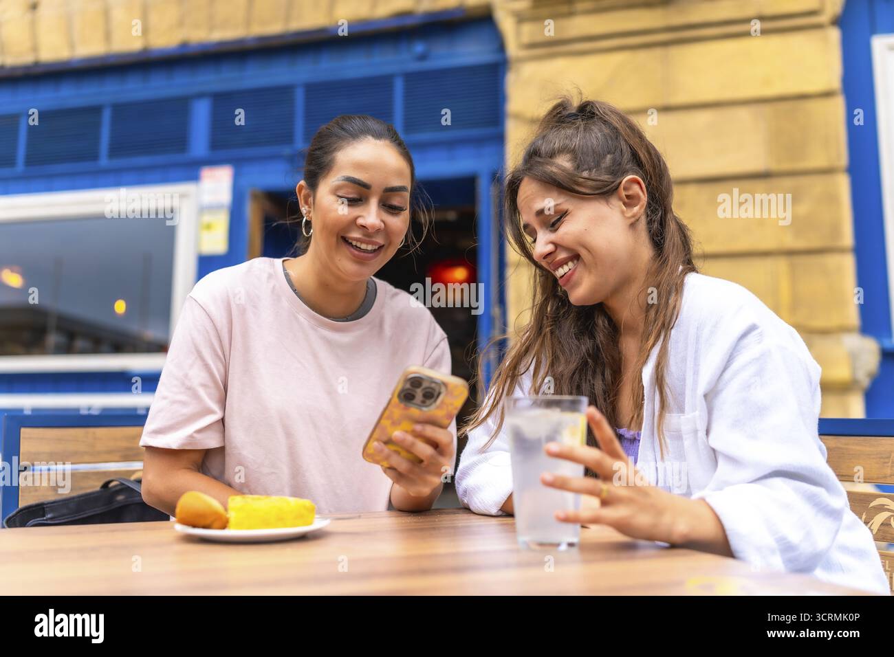 Due amiche si stanno godendo un momento insieme in un bar, guardando qualcosa di divertente su uno smartphone e ridendo Foto Stock