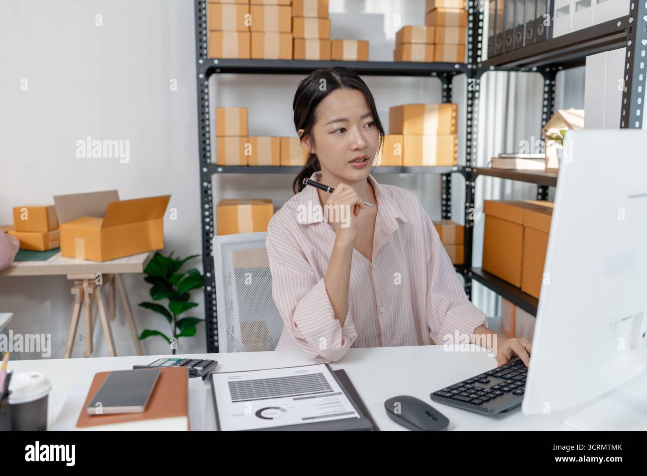 Una giovane donna con una camicia a righe lavora alla sua scrivania con documenti, calcolatrice e smartphone, concentrandosi sulle attività contabili e sulla pianificazione finanziaria Foto Stock