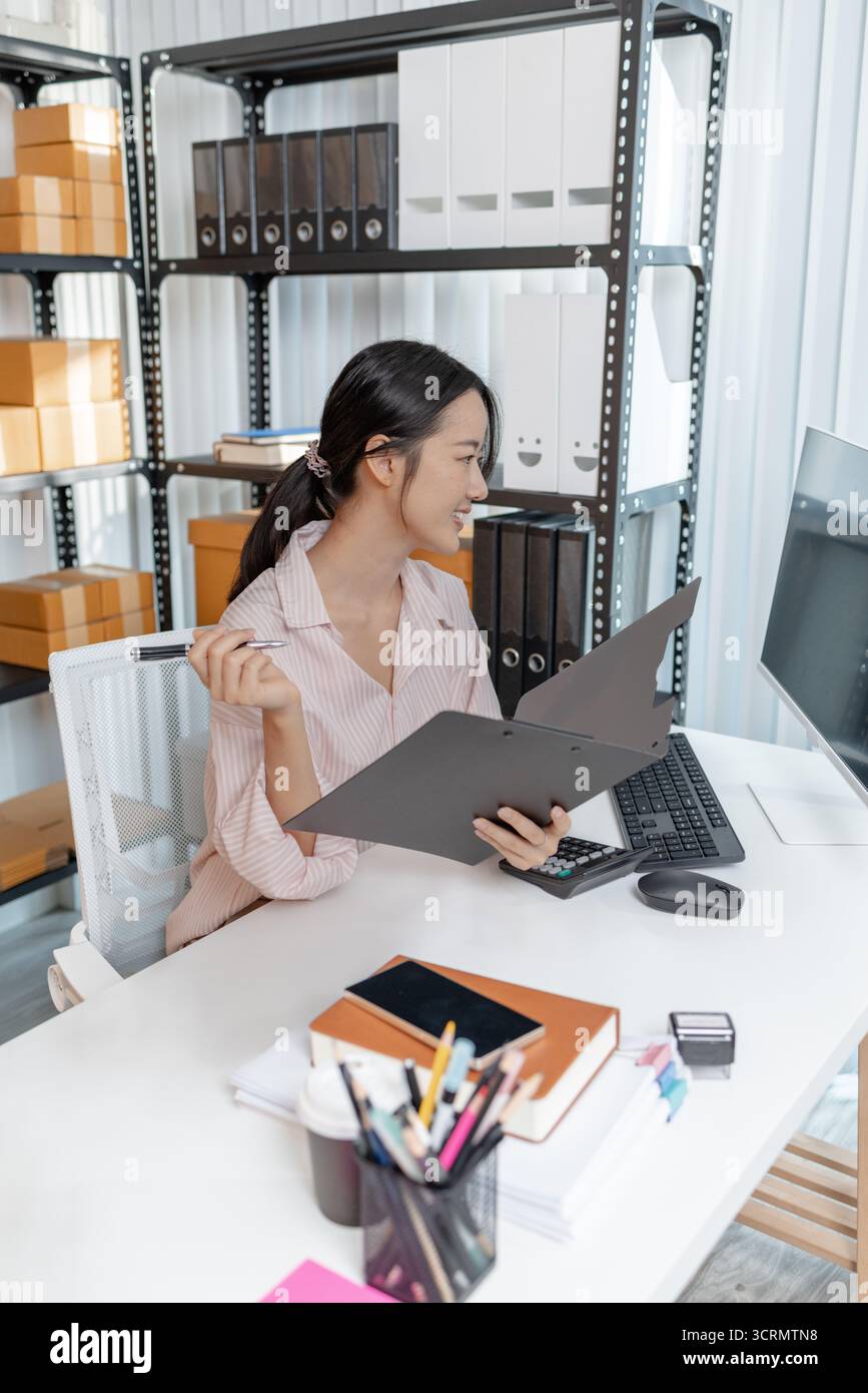 Una giovane donna con una camicia a righe lavora alla sua scrivania con documenti, calcolatrice e smartphone, concentrandosi sulle attività contabili e sulla pianificazione finanziaria Foto Stock