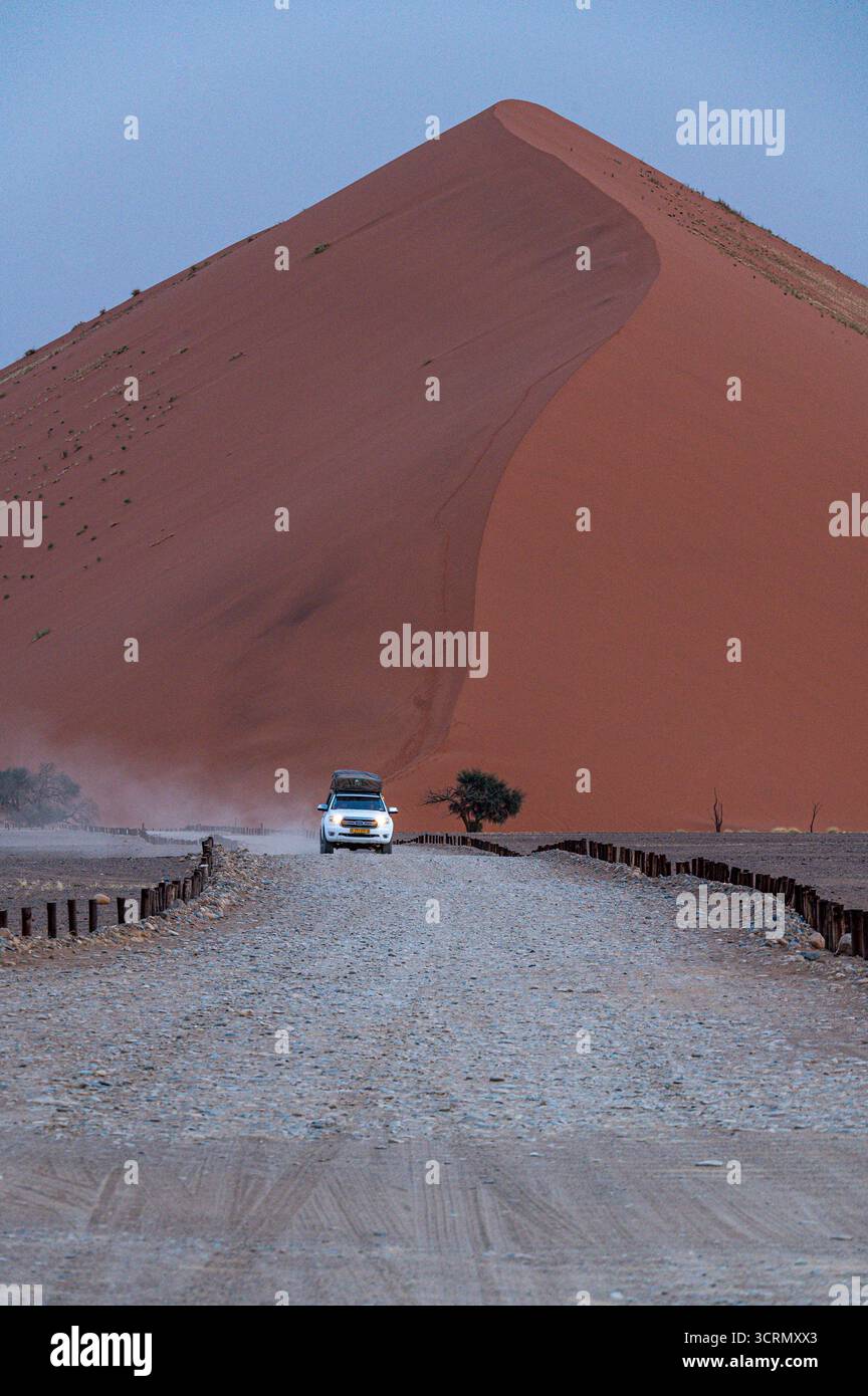 Vista dall'angolo basso di un veicolo 4x4 solitario che guida una strada desertica ghiaia verso una torreggiante duna arancione-ruggine, evocando avventura, Namib-Naukluft, Namibia. Foto Stock