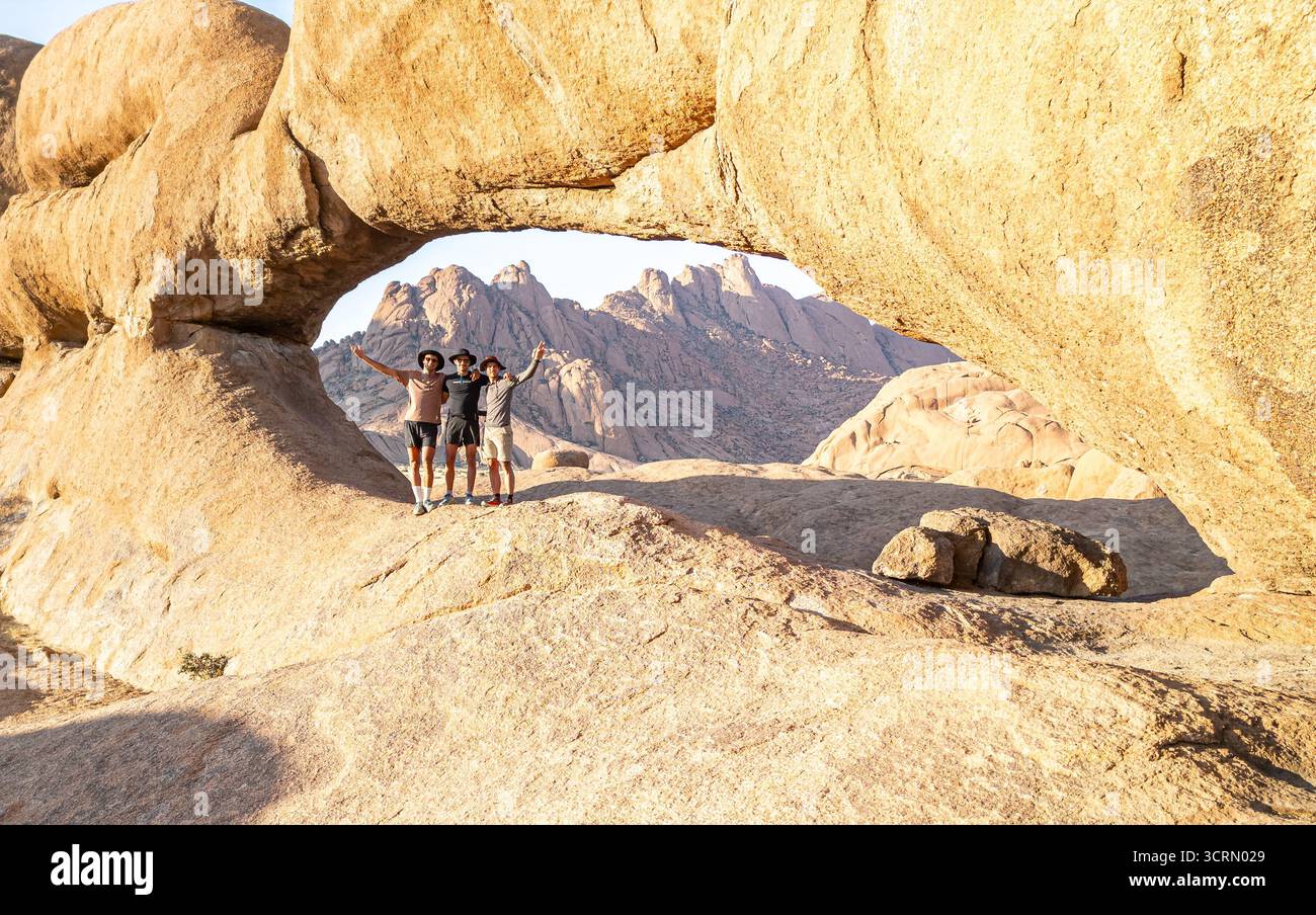 Tre escursionisti si pongono gioiosamente sotto l'iconico arco di roccia granitica di Spitzkoppe, le suggestive vette di inselberg visibili attraverso l'apertura, la regione di Erongo, Namibia. Foto Stock