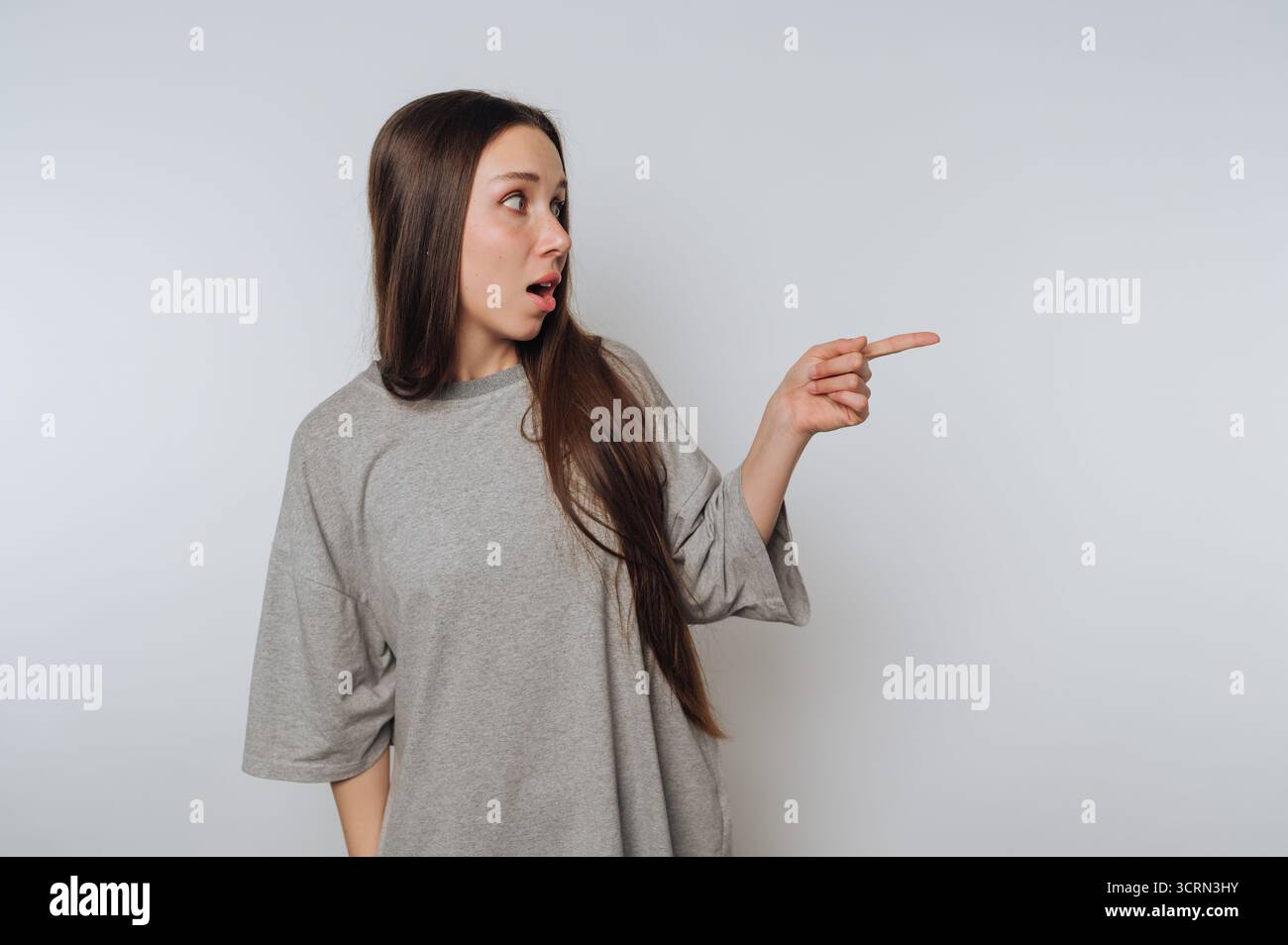 Una giovane donna dai capelli lunghi che indossa una camicia oversize grigia mostra sorpresa e punta di lato. Appare impegnata ed espressiva in un minimalista Foto Stock