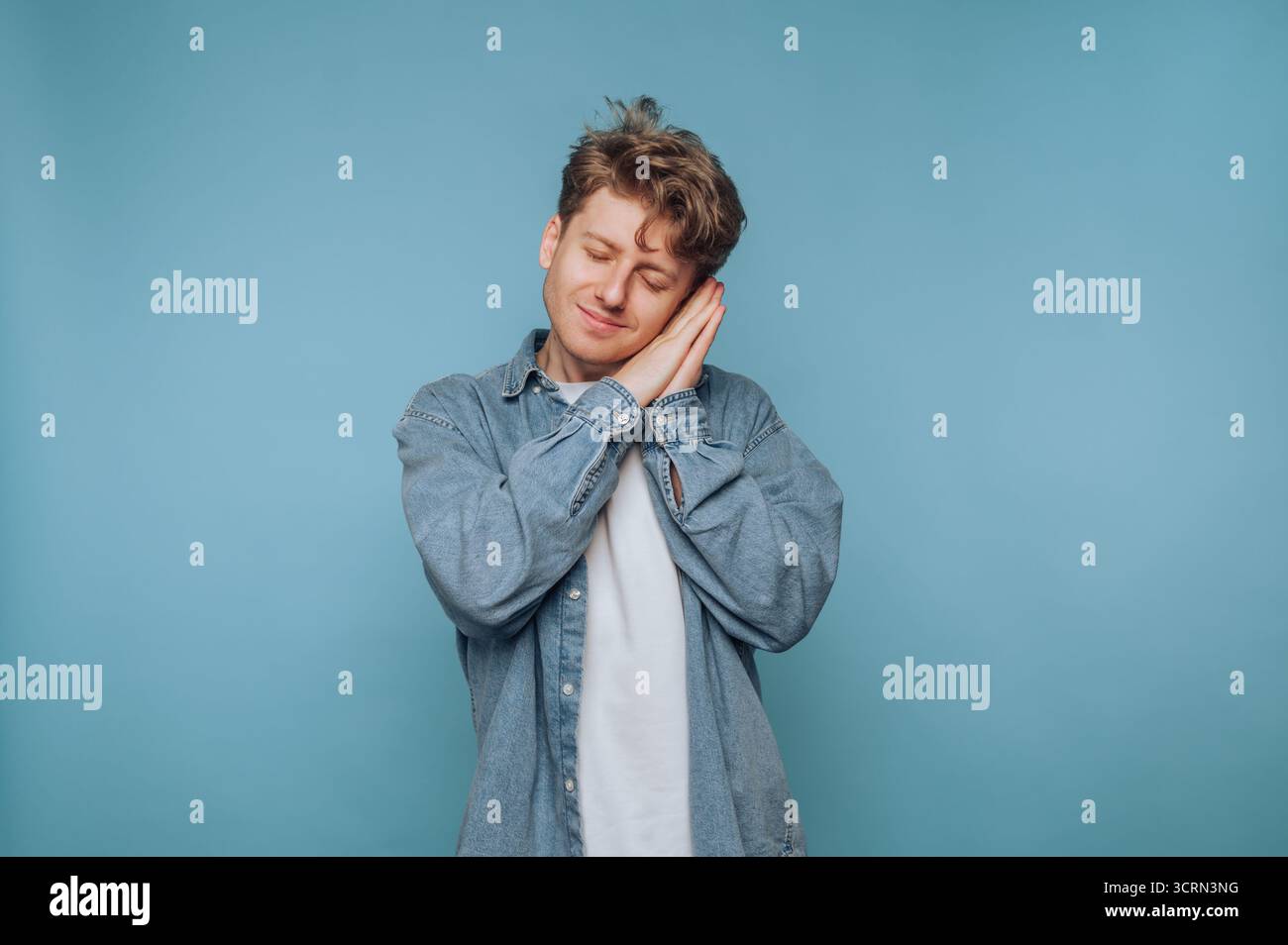 Un giovane con i capelli ricci sorride mentre simula una posa addormentata, mostrando un umore gioioso. Il morbido sfondo blu migliora l'atmosfera giocosa, Foto Stock