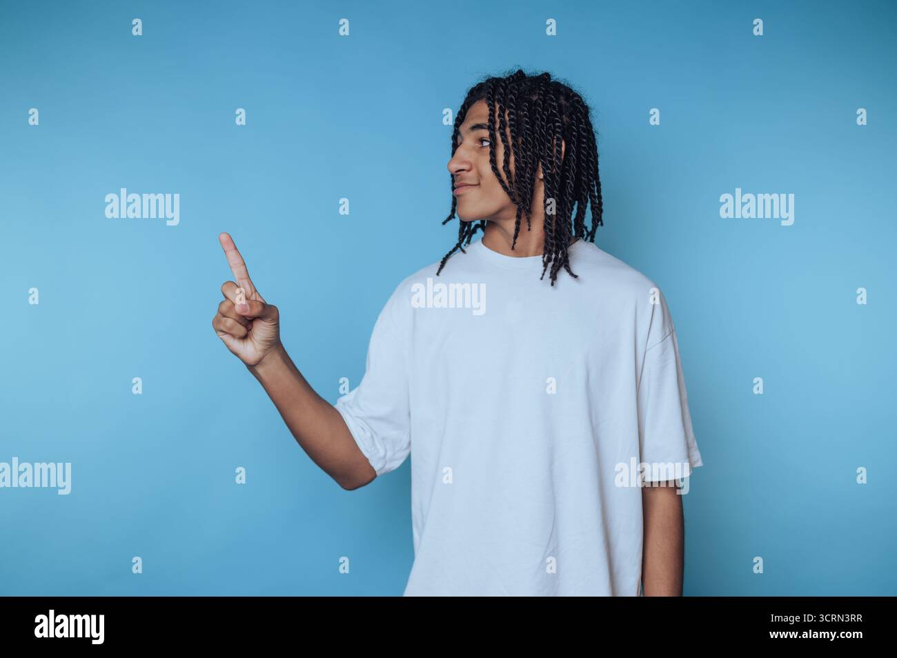 Un giovane con i capelli intrecciati punta giocosamente il dito mentre sorride su un solido sfondo blu in un ambiente creativo o informale. Il suo espresso Foto Stock