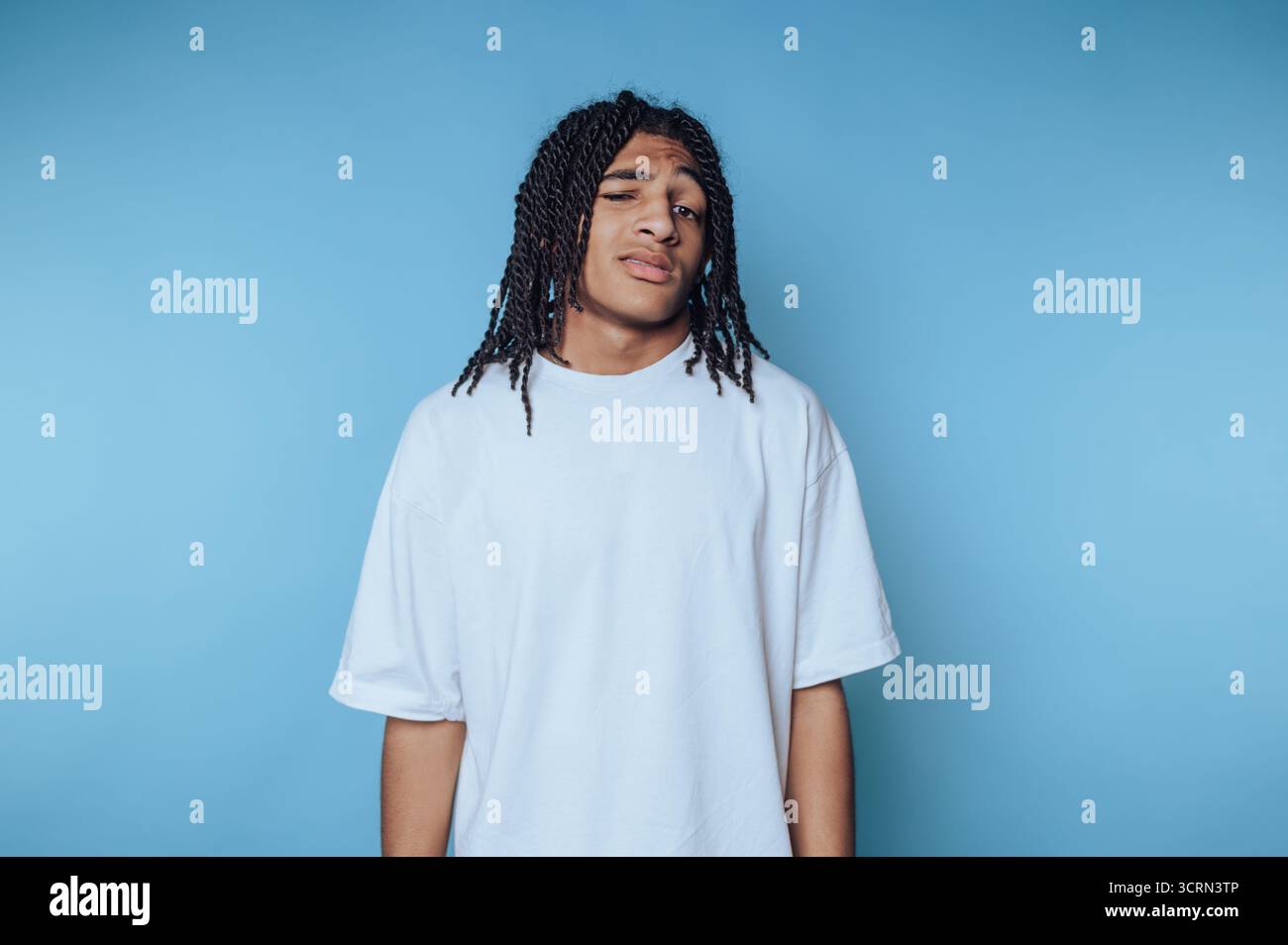 Un giovane uomo è in piedi con una postura rilassata e indossa una semplice t-shirt bianca. La sua acconciatura intrecciata completa la sua espressione premurosa contro un Foto Stock