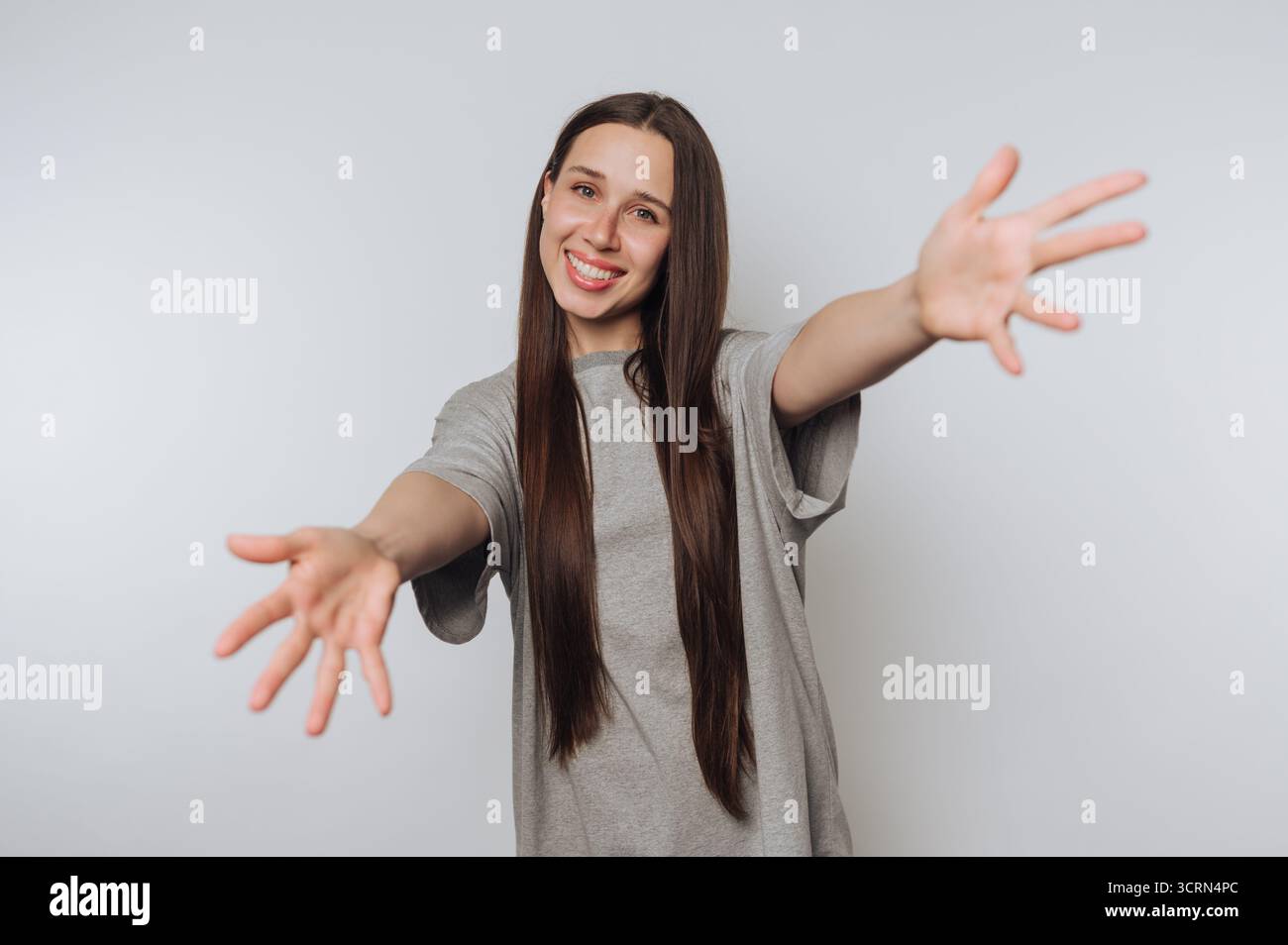Una giovane donna con lunghi capelli marroni sorridente e che si allontana a braccia aperte, indossa una t-shirt grigia casual su un semplice sfondo bianco. Foto Stock