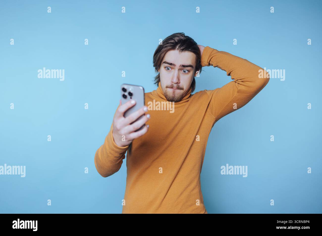 ritratto di un giovane uomo che tiene in mano un telefono mentre mostra preoccupazione, cattura le sfumature della vita digitale moderna su sfondo blu Foto Stock