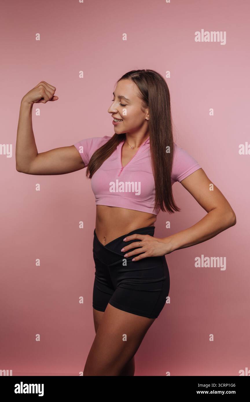 Una donna sta in piedi con fiducia, piegando il braccio per mostrare i muscoli mentre sorride. Indossa un top rosa e pantaloncini neri, con una vivace b rosa Foto Stock