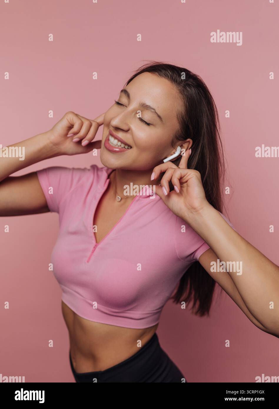Una giovane donna con i capelli lunghi, che indossa una cover rosa, sorride mentre ascolta la musica con gli auricolari wireless. L'espressione vivace trasmette il suo godimento Foto Stock