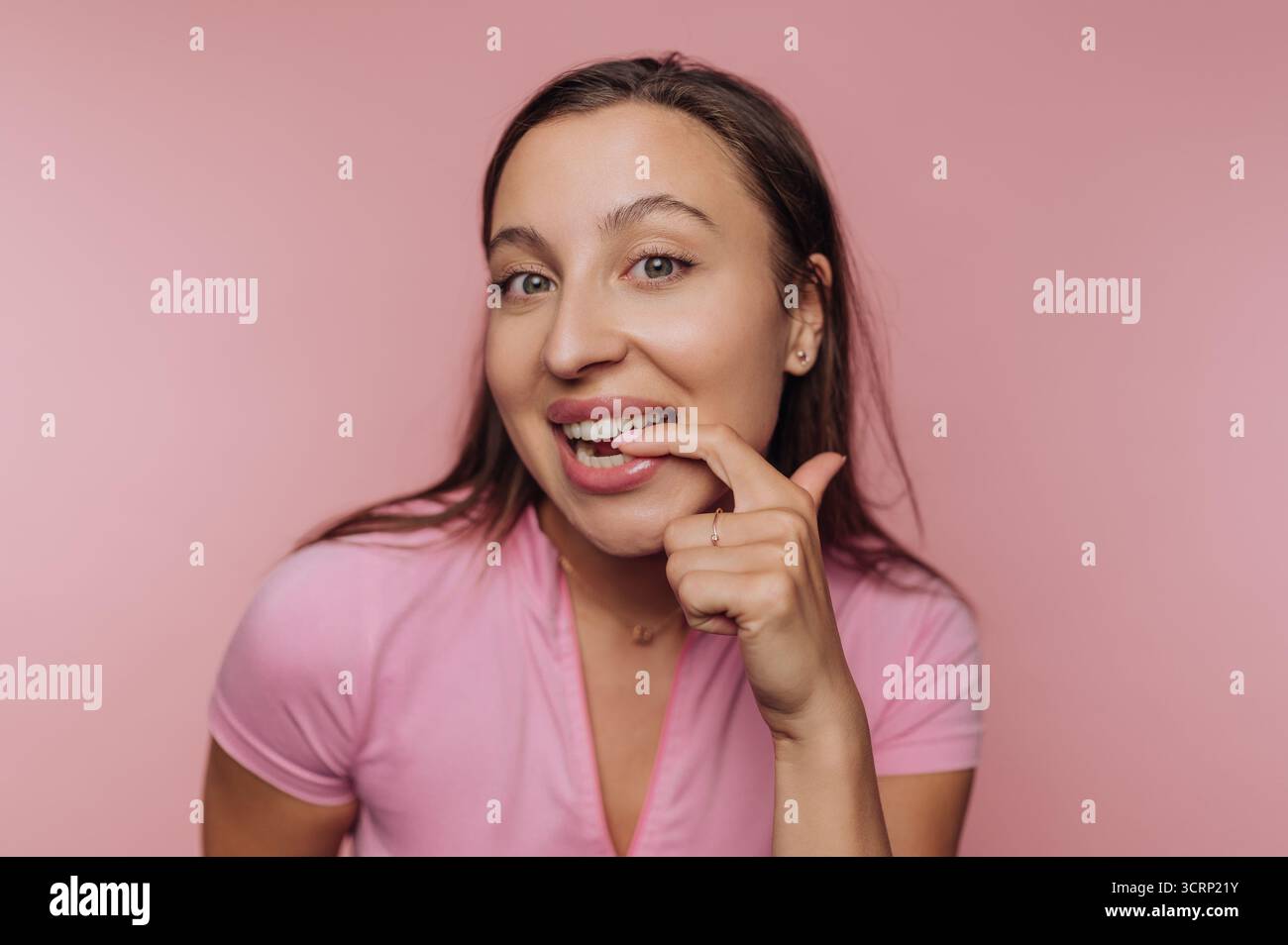 Una donna dai capelli lunghi mostra un'espressione gioiosa toccando leggermente i denti con un dito. Indossa un top rosa in una vivace stanza rosa. Foto Stock