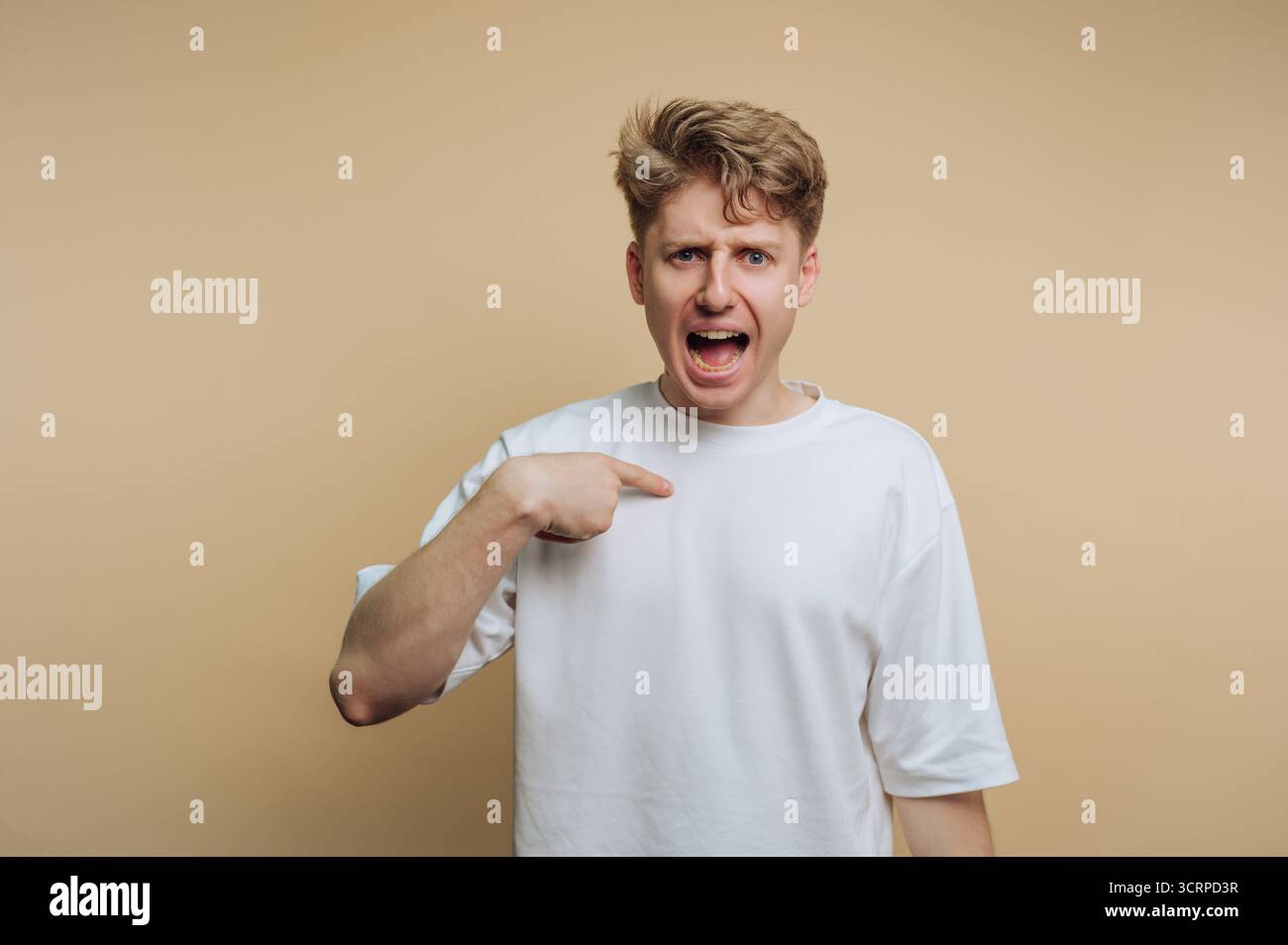 Un uomo con i capelli ruvidi appare animato mentre si indica a se stesso, mostrando le sue emozioni. Lo sfondo neutrale enfatizza i suoi gesti espressivi, Foto Stock