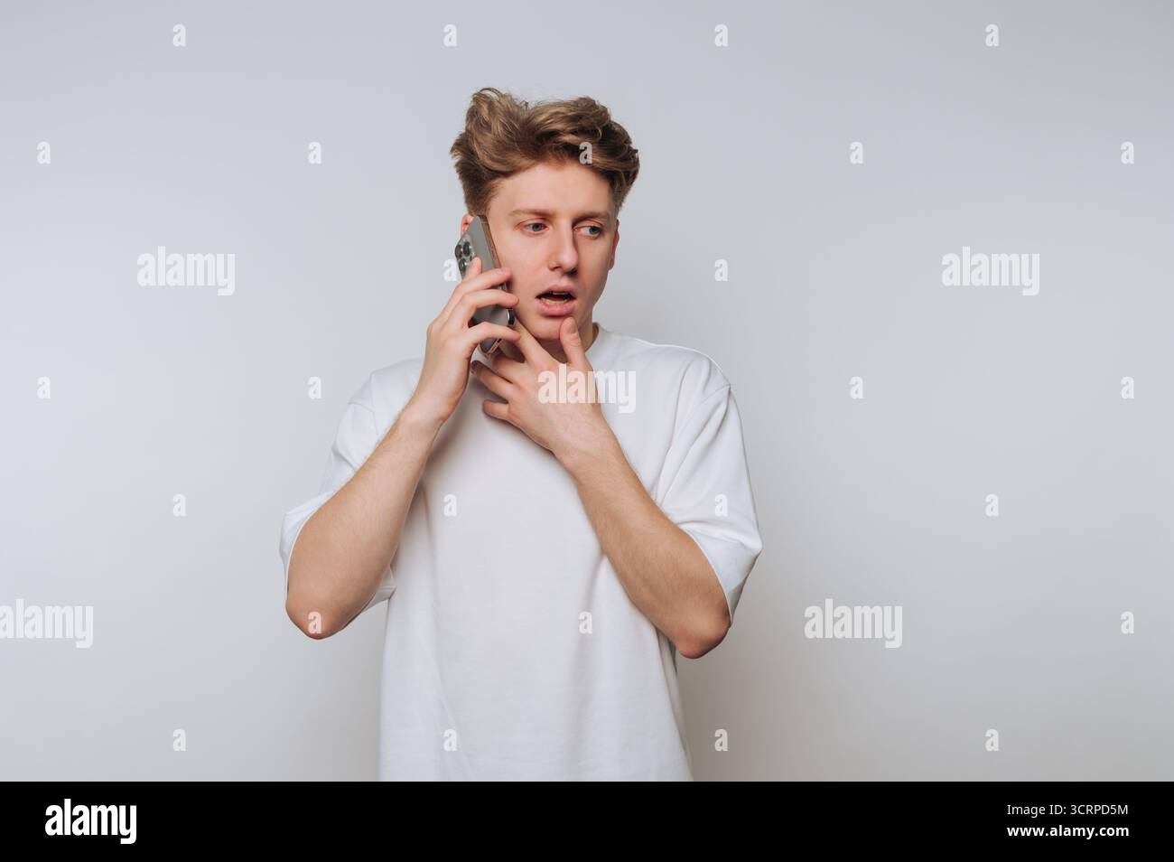 Colto in un momento di sorpresa, un giovane con una t-shirt bianca tiene uno smartphone all'orecchio, mostrando segni di preoccupazione mentre si trova in piedi contro un pla Foto Stock