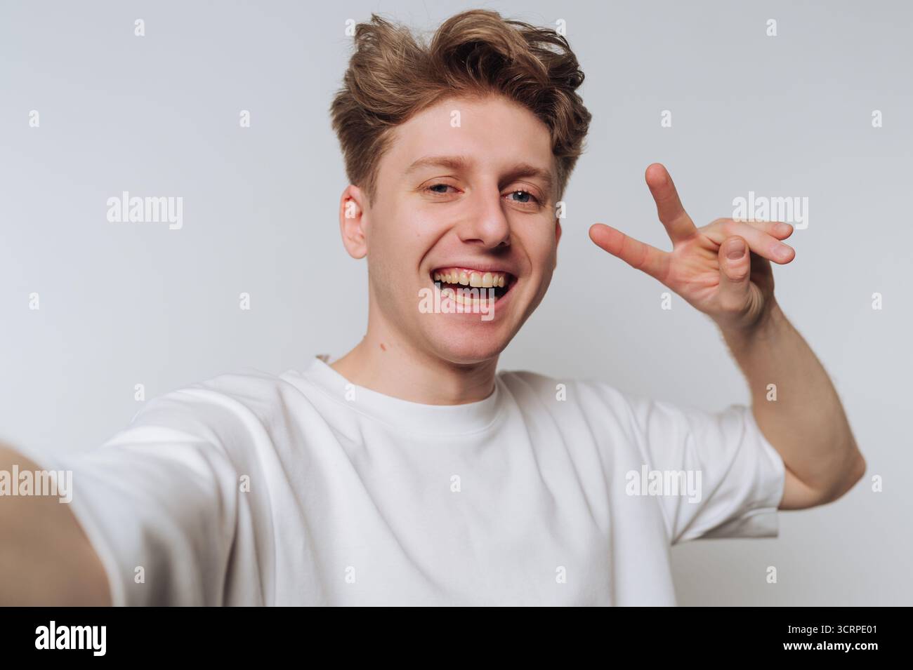 Un giovane uomo si trova su uno sfondo semplice, raggiante di gioia mentre fa un selfie. Lancia un segno di pace con una mano, sembrando amichevole e allegro Foto Stock