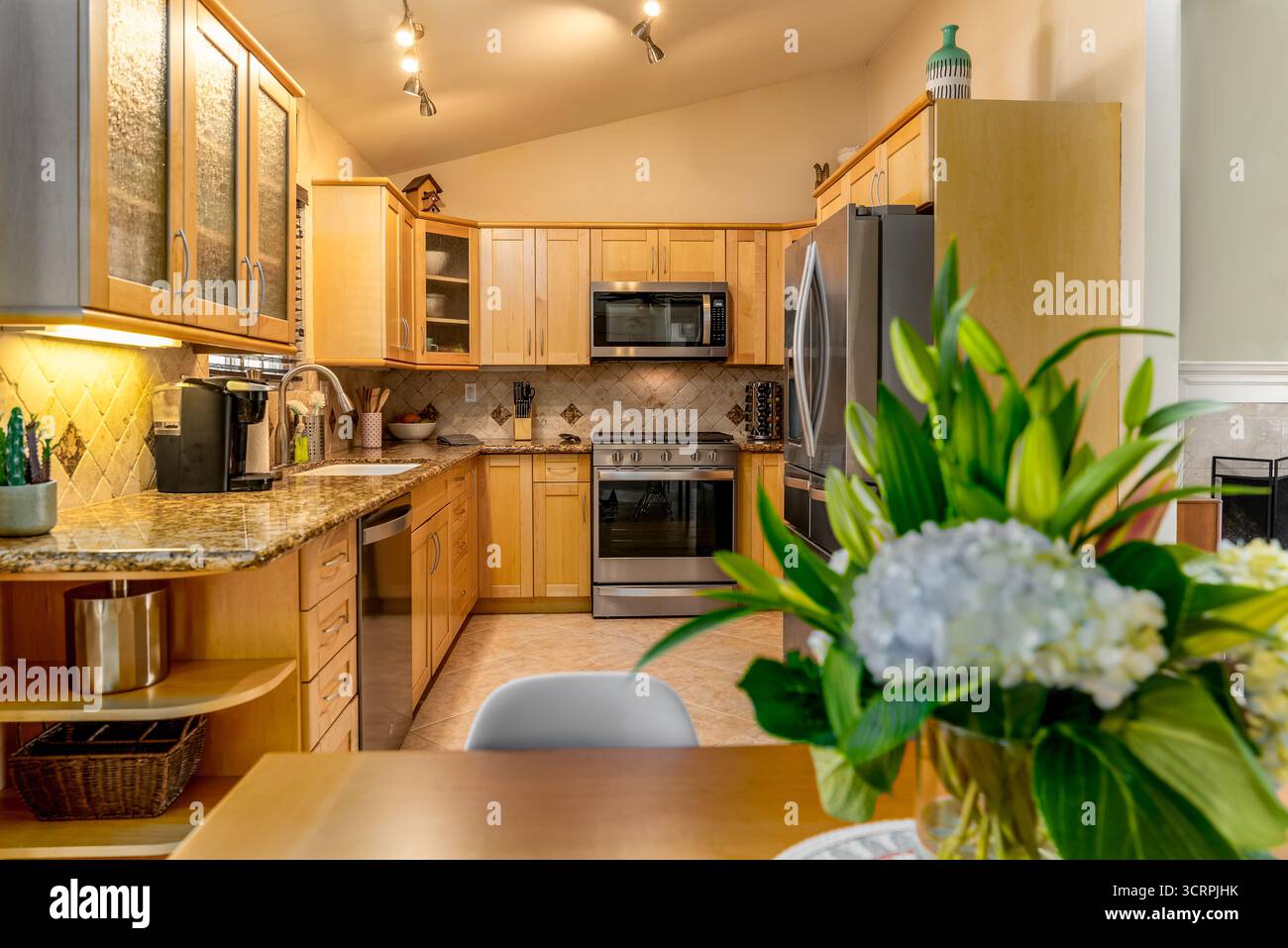 Cucina armadi in legno naturale ripiani in granito elettrodomestici in acciaio inox tavolo da pranzo bouquet di fiori in primo piano Foto Stock
