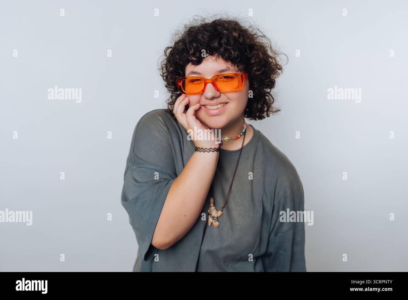 Persona sorridente con capelli ricci che indossa occhiali da sole arancioni e una t-shirt grigia. Foto Stock
