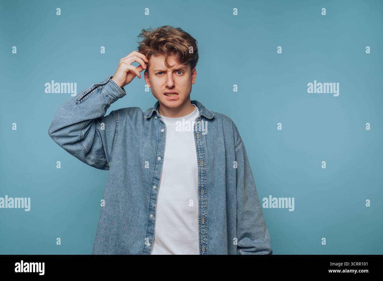 Un giovane si trova di fronte a un solido sfondo blu, sembra sconcertato e graffiato la testa, trasmettendo un senso di confusione e di profonda riflessione Foto Stock