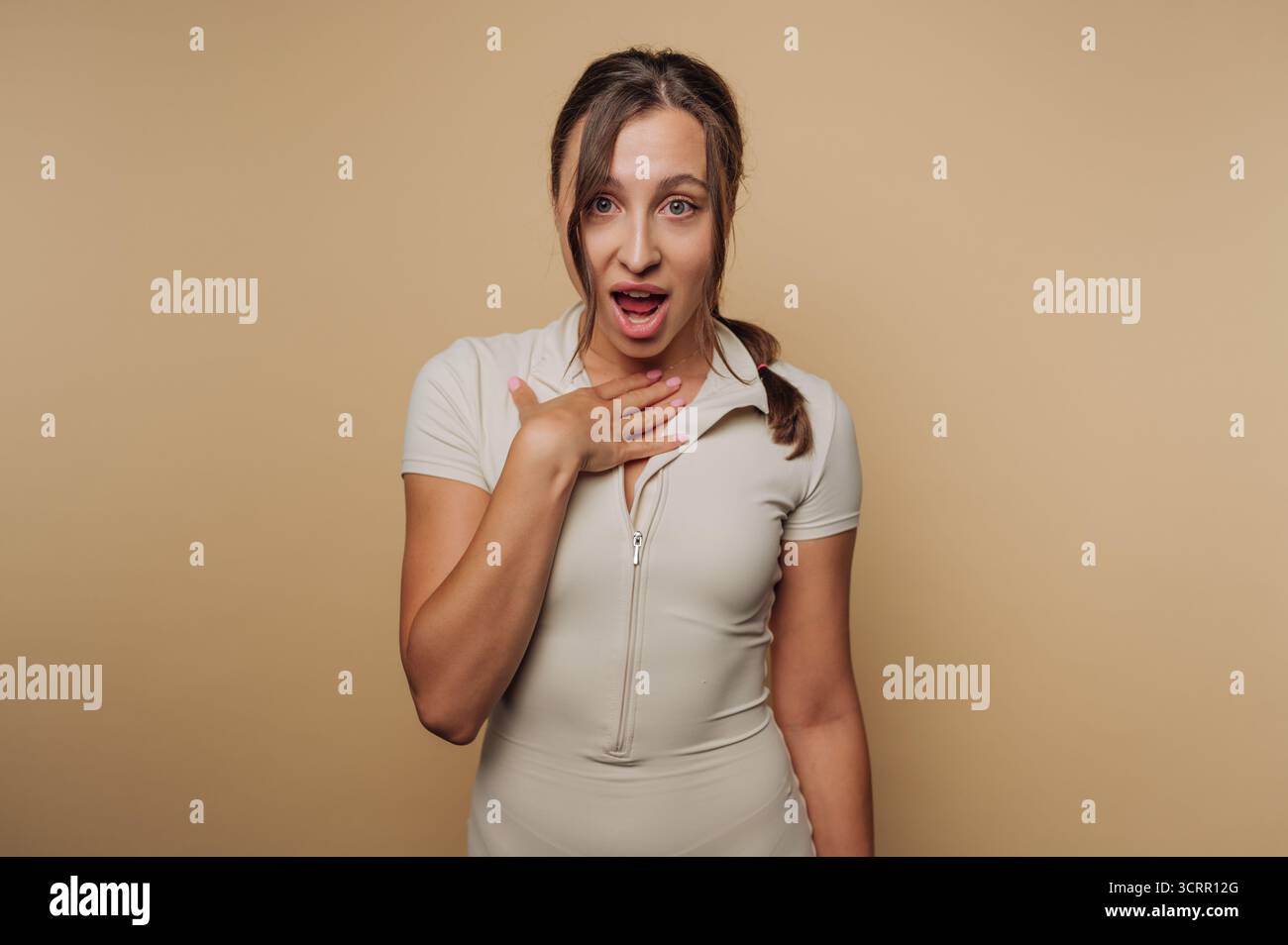 Una donna è colta in un momento di sorpresa, mano posta sul petto e bocca leggermente aperta. Lo sfondo dai toni neutri enfatizza la sua espressione An Foto Stock