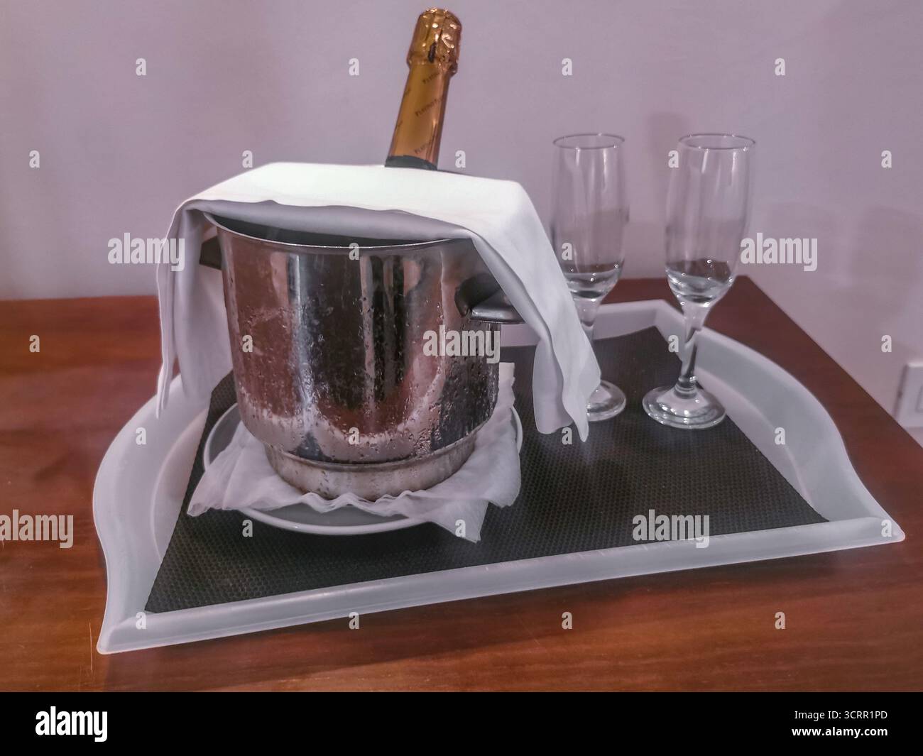 Romantica sistemazione di benvenuto per la luna di miele in una camera d'hotel: Champagne ghiacciato in un secchio di ghiaccio d'argento avvolto in un tovagliolo bianco, accompagnato da due eleganti vetri Foto Stock