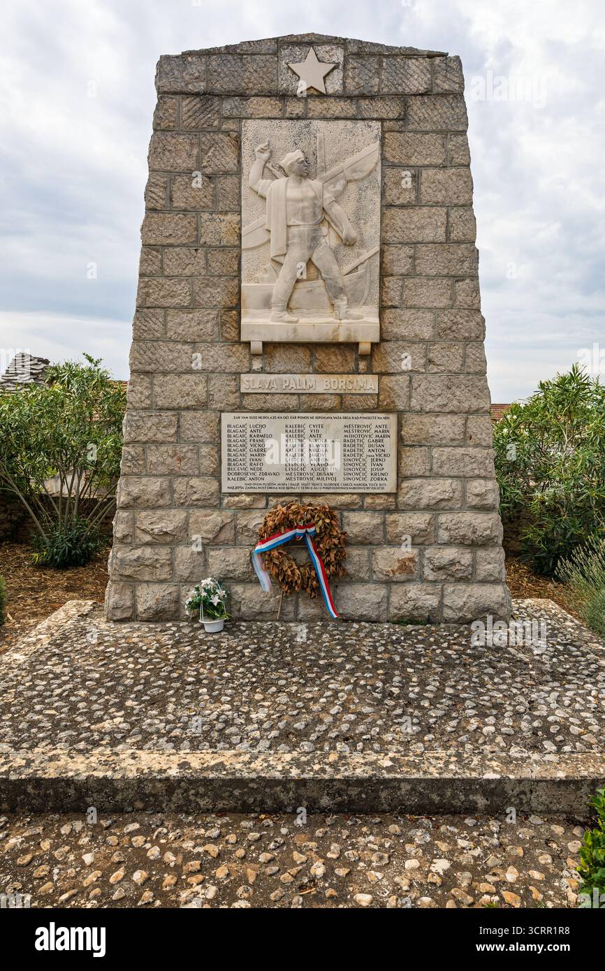 DONJE SELO, CROAZIA, 08.24.2025: Un solenne memoriale di guerra in pietra presenta una figura operaia scolpita e un emblema della stella, che commemora gli eroi caduti. Una corona r Foto Stock