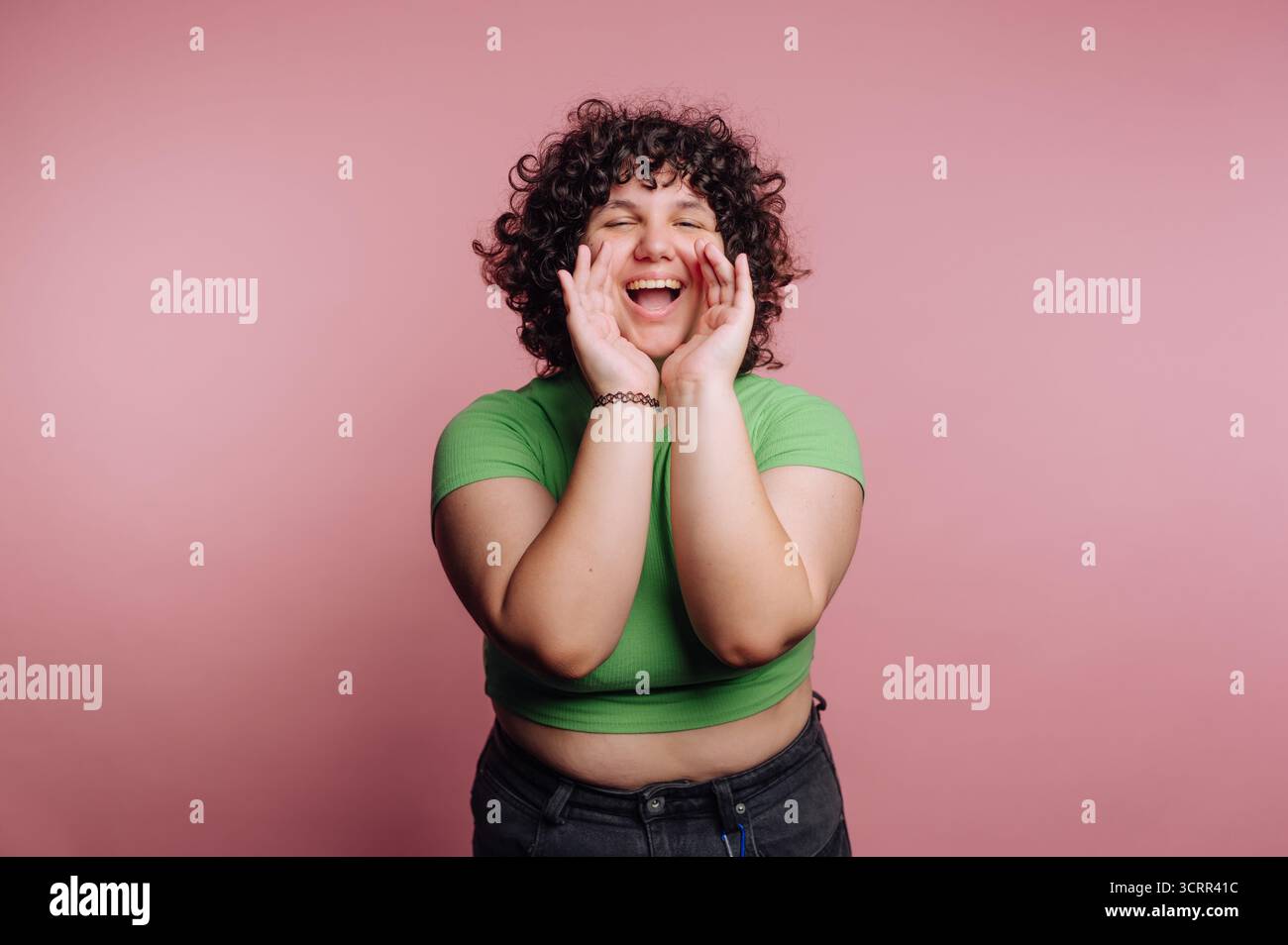 Una donna con i capelli ricci sorride brillantemente e gioca a posare con le mani per il viso. Lo sfondo rosa brillante esalta l'atmosfera gioiosa. C Foto Stock