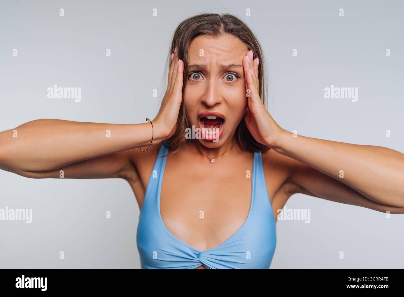 Una donna esprime shock e sorpresa intensi con gli occhi larghi e la bocca aperta. Tiene la testa con entrambe le mani, trasmettendo forti emozioni in un neut Foto Stock