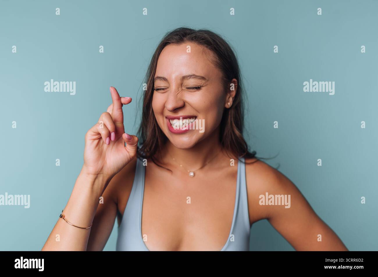 Una giovane donna sta in piedi con fiducia, sorridendo e giocherellando le dita. Ha i capelli a spalla e indossa una canotta azzurra. Il backgr Foto Stock Una giovane donna sta in piedi con fiducia, sorridendo e giocherellando le dita. Ha i capelli a spalla e indossa una canotta azzurra. Il backgr Foto Stock