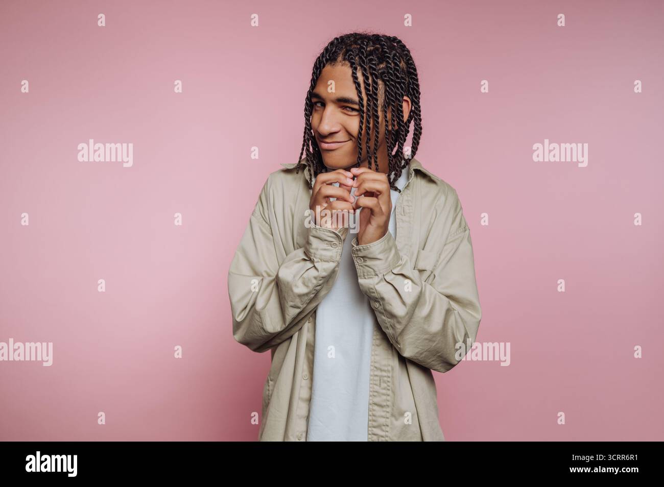 Un giovane si trova su uno sfondo rosa tenue, grattando e tenendo le mani giocosamente vicino al viso. I suoi capelli intrecciati e la sua camicia rilassata riflettono Foto Stock