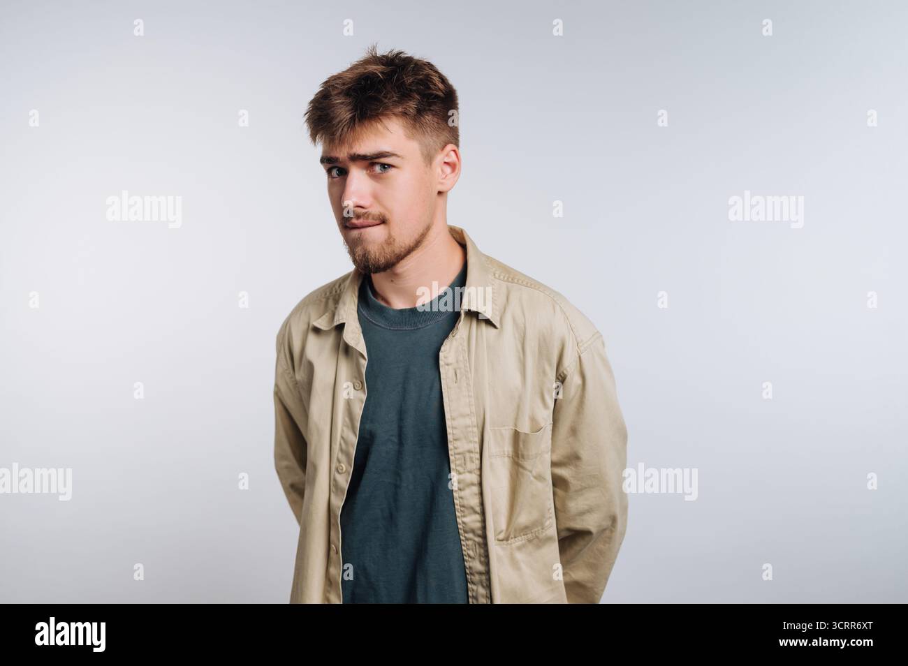 Un giovane sta con le mani dietro la schiena in una posa informale. Ha i capelli corti e la barba, indossa una giacca di colore chiaro su una t-shirt. Foto Stock
