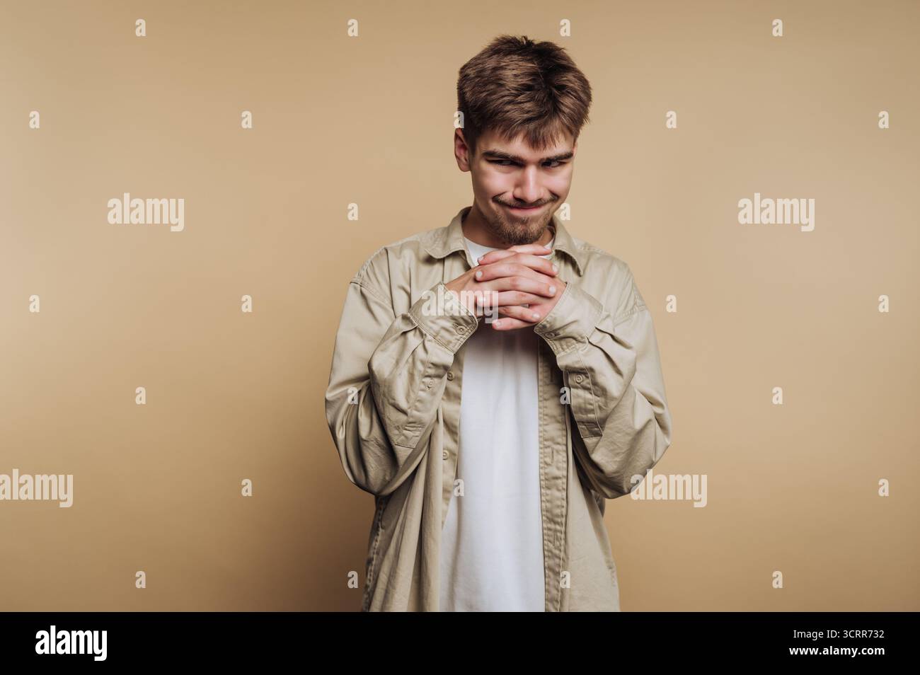 Un giovane sta con un sorriso sottile, tenendo le mani unite. È vestito con una camicia leggera e sembra essere di umore riflessivo. Il backgro Foto Stock