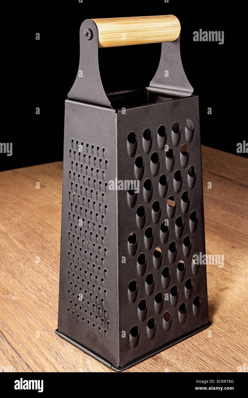 Questa immagine presenta un'elegante grattugia nera progettata per tritare le verdure. Poggia su un rustico tavolo da cucina in legno, sottolineandone la versatilità e la versatilità Foto Stock