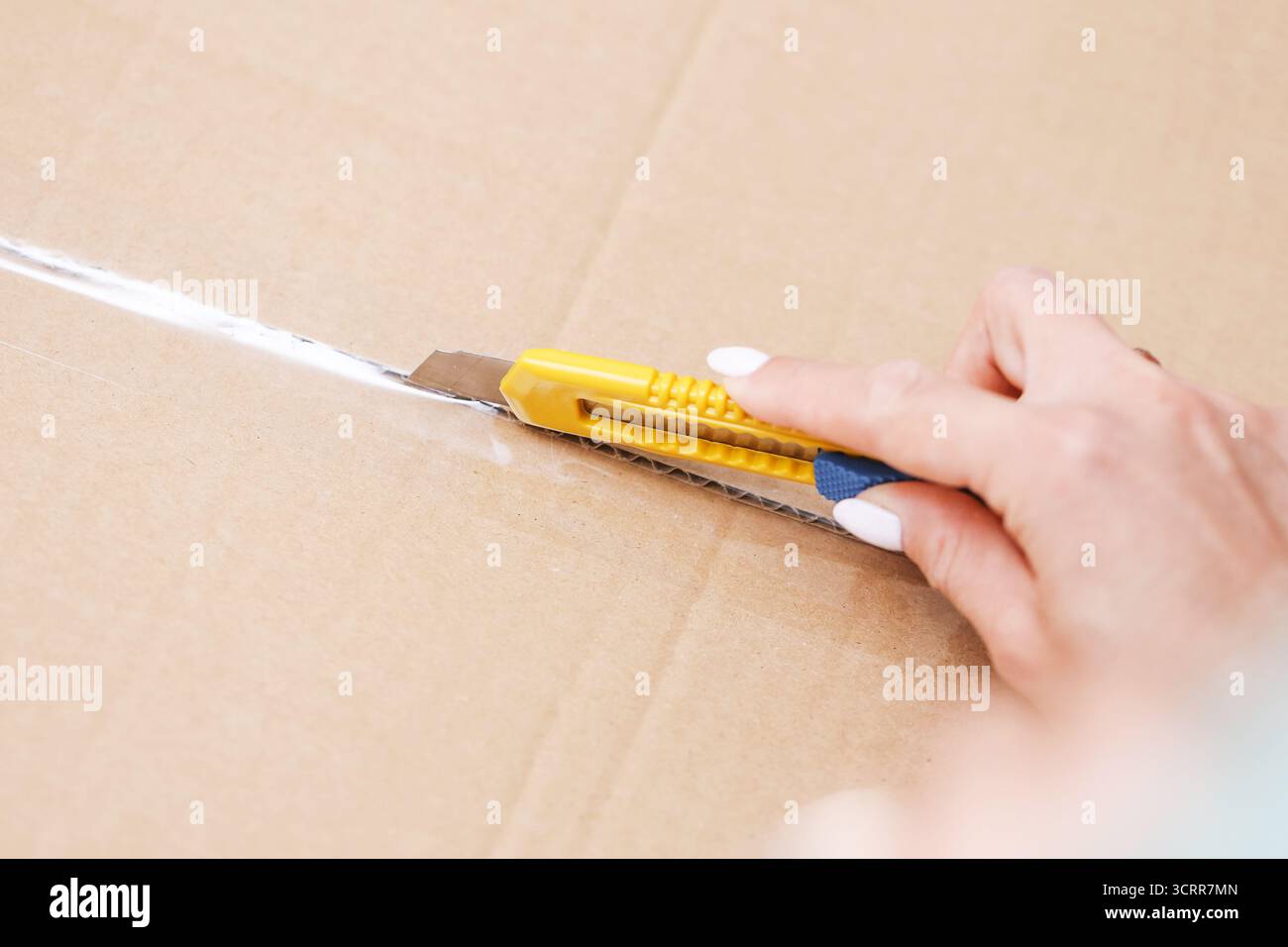 Tagliare a mano la scatola di cartone utilizzando un coltello giallo. Processo di imballaggio e disimballaggio a casa. Concetto di famiglia o di consegna. Tecnica di taglio sicura. Foto Stock