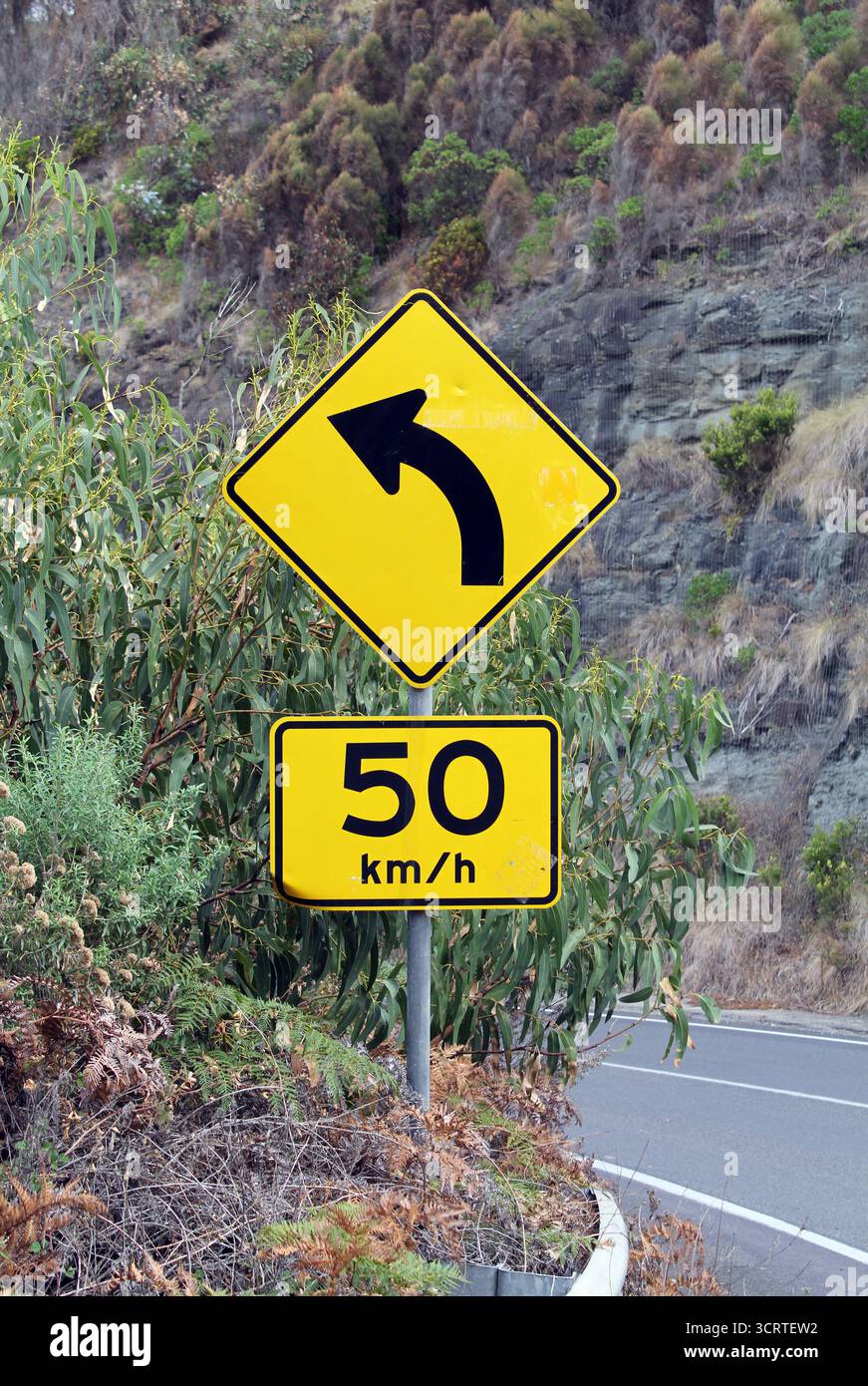 Limite di velocità giallo e svolta a sinistra tra gli alberi e una scogliera Foto Stock