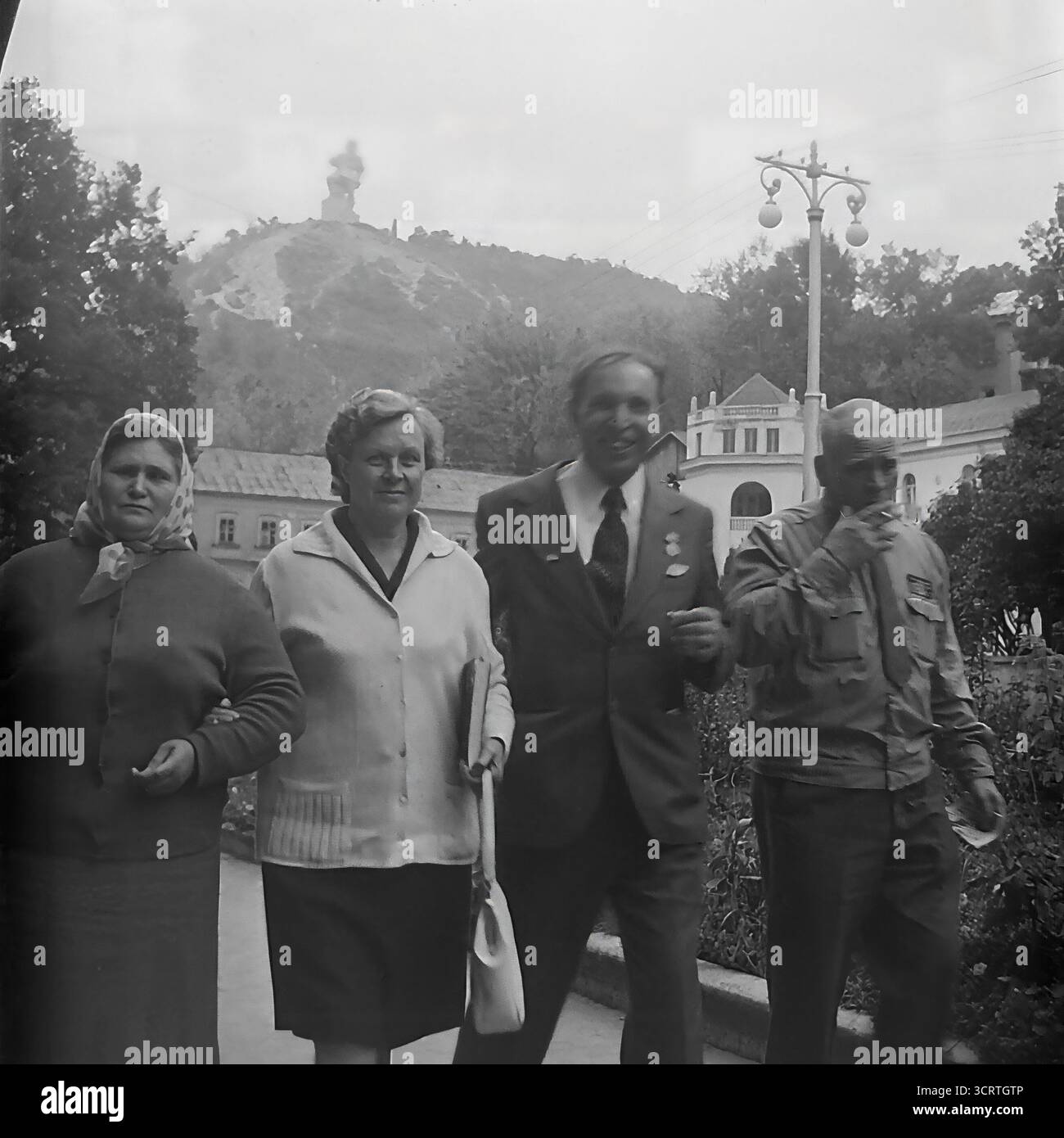 Una candida fotografia d'archivio in bianco e nero degli anni '1970, che mostra un gruppo di cittadini sovietici che si godono una passeggiata presso la stazione termale di Sviatohirsk, la RSS Ucraina. Il gruppo, che include un veterano di guerra decorato, sta passeggiando attraverso i giardini del parco. Sullo sfondo, il famoso Monumento colossale ad Artem è chiaramente visibile in cima a una collina di gesso, che si affaccia sul resort. Questa immagine nostalgica cattura un momento di tranquillo svago e turismo in una popolare città termale di epoca sovietica, con un importante punto di riferimento ideologico che forma lo sfondo della vita quotidiana Foto Stock